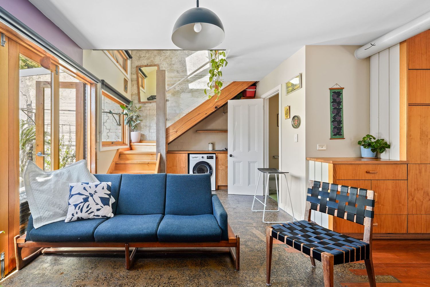 Modern living room interior with blue sofa, wooden staircase, and natural light from large windows.