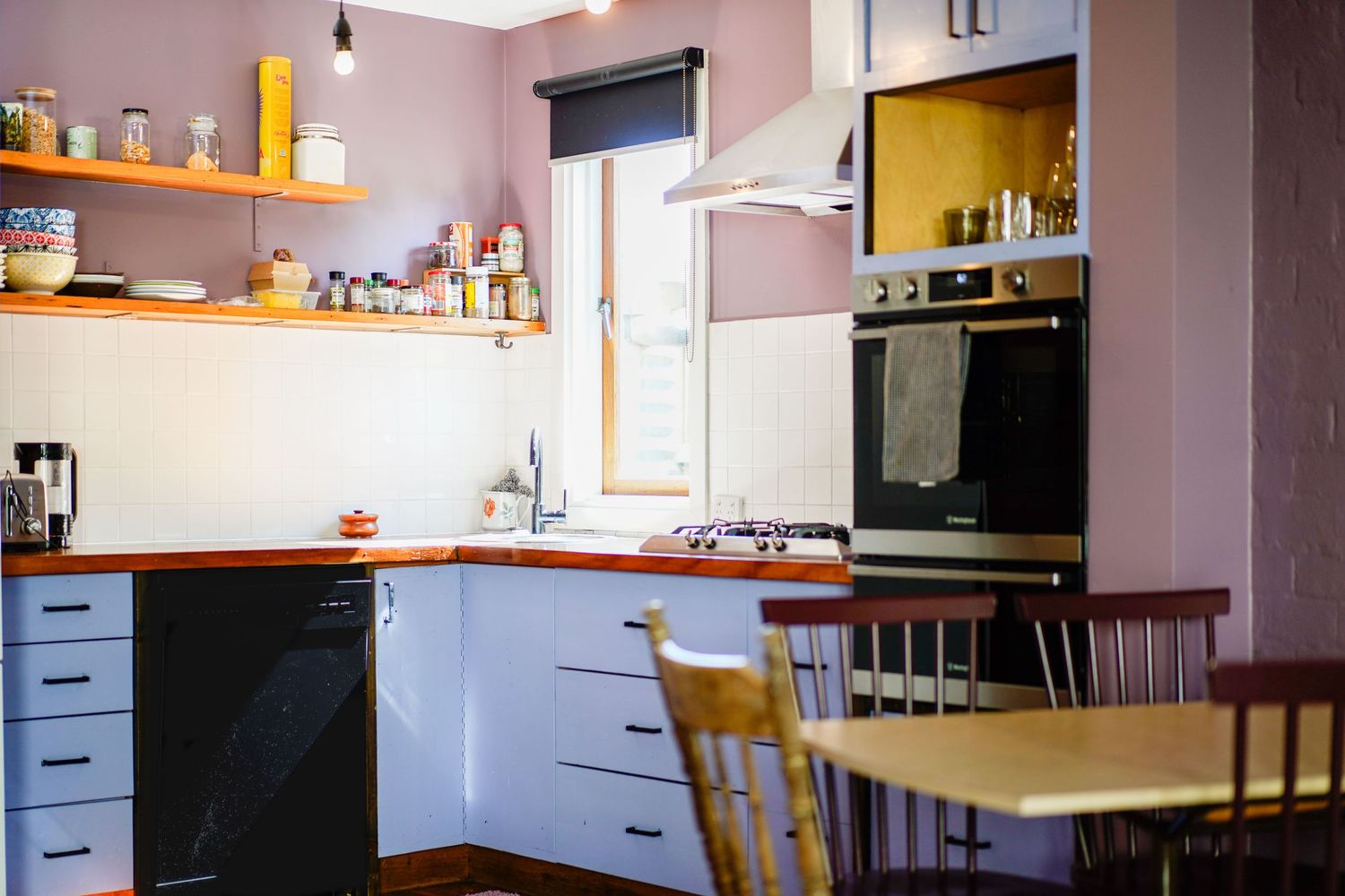 Modern corner kitchen with pink walls, white cabinets, wooden shelving and dining furniture in natural light.
