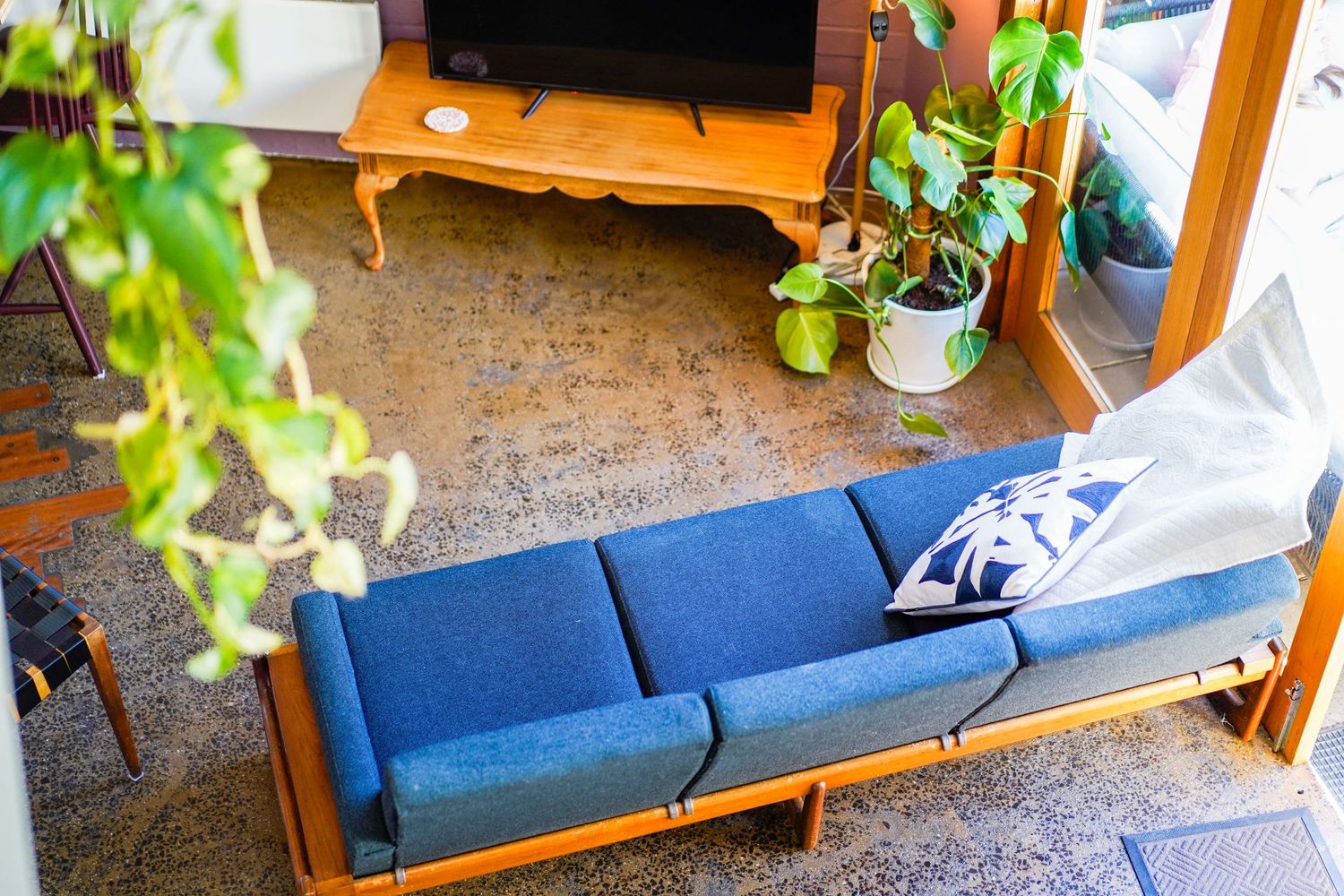 Mid-century modern blue couch with wooden frame sits in a living room corner beside plants and TV stand.