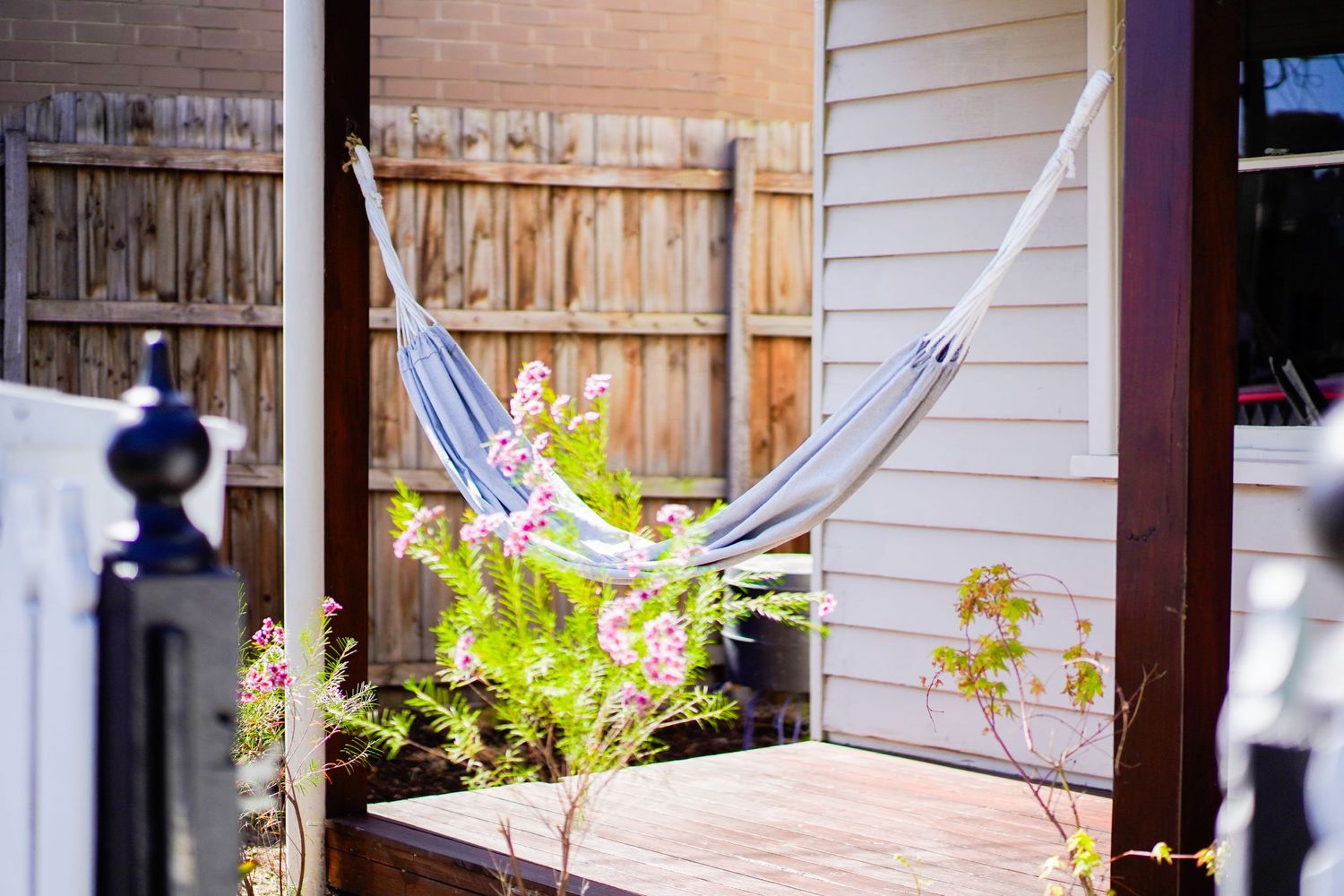 A hammock hangs between wooden posts on a sunny porch with flowers and a wooden fence in the background.