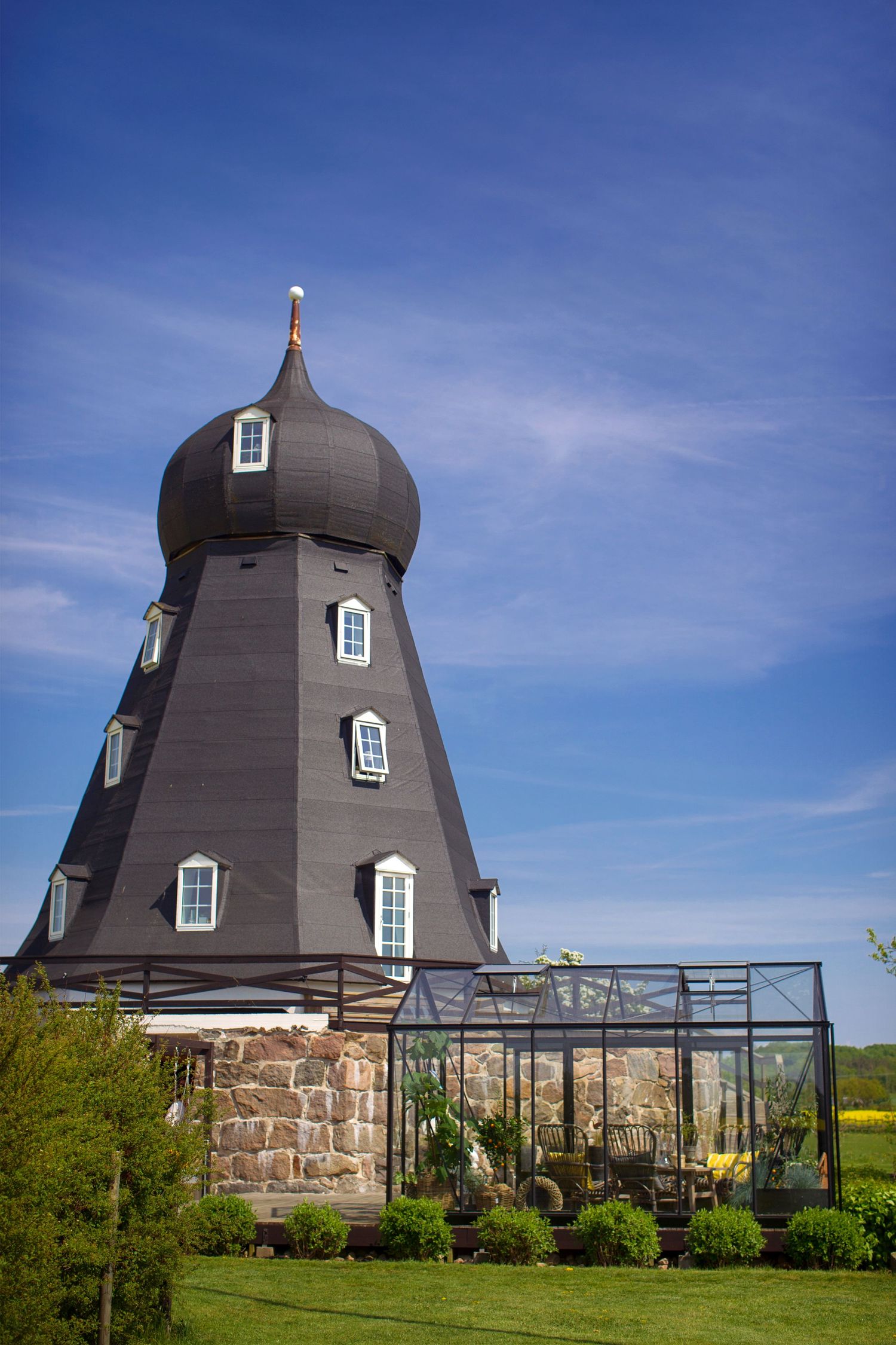 Unique windmill-style building with a dark onion dome and small windows next to a greenhouse structure under blue sky.