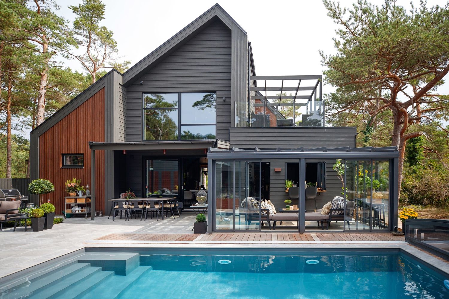 Modern house exterior with swimming pool and spacious deck area surrounded by pine trees.