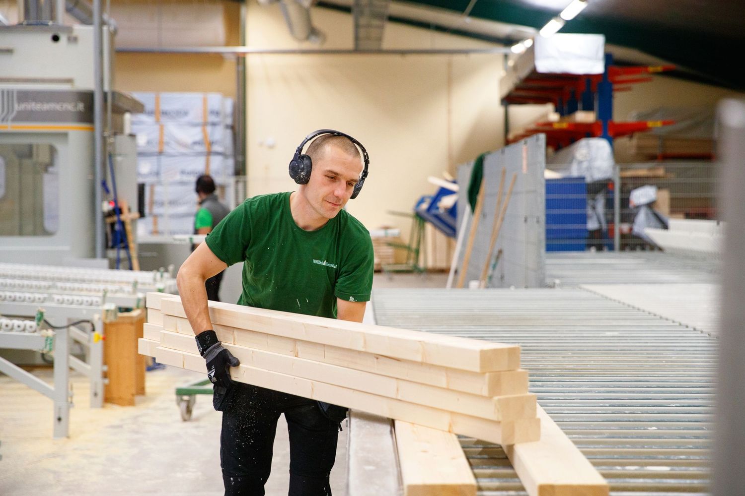 Worker carrying lumber while wearing protective headphones in an industrial workshop.