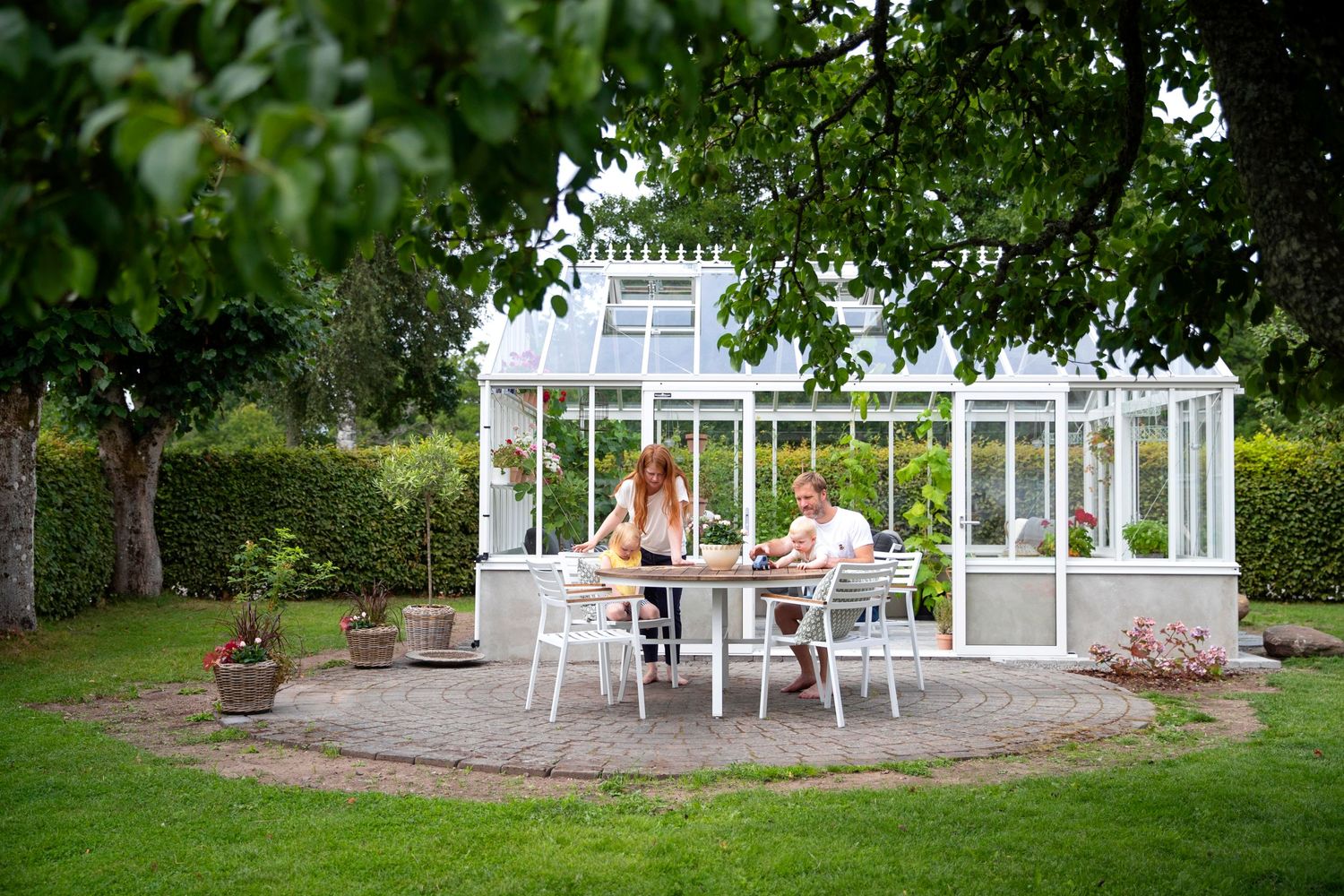 White greenhouse conservatory in a garden setting with outdoor dining furniture.