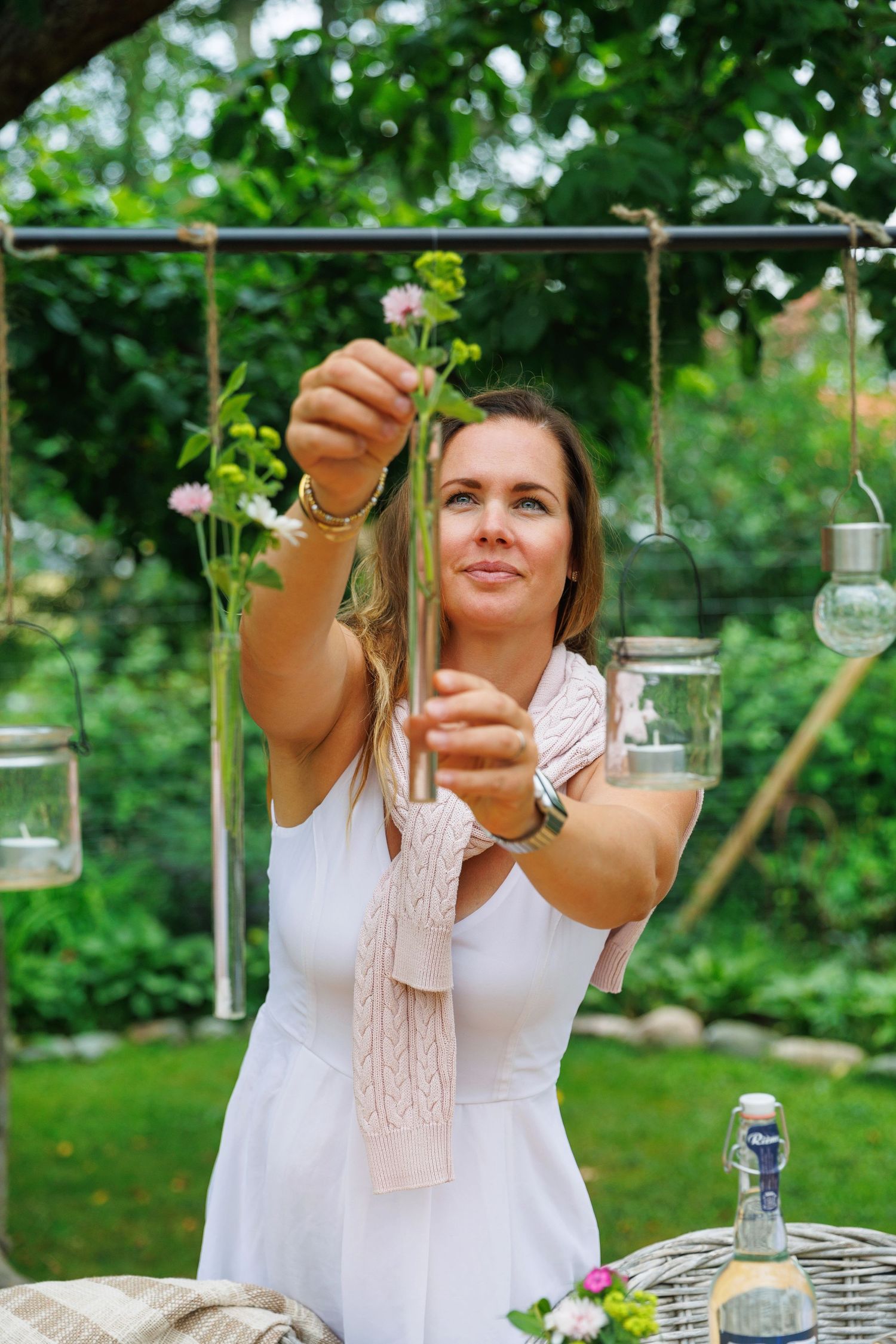 Woman in a white dress decorating an outdoor garden space with hanging glass lanterns and flowers.
