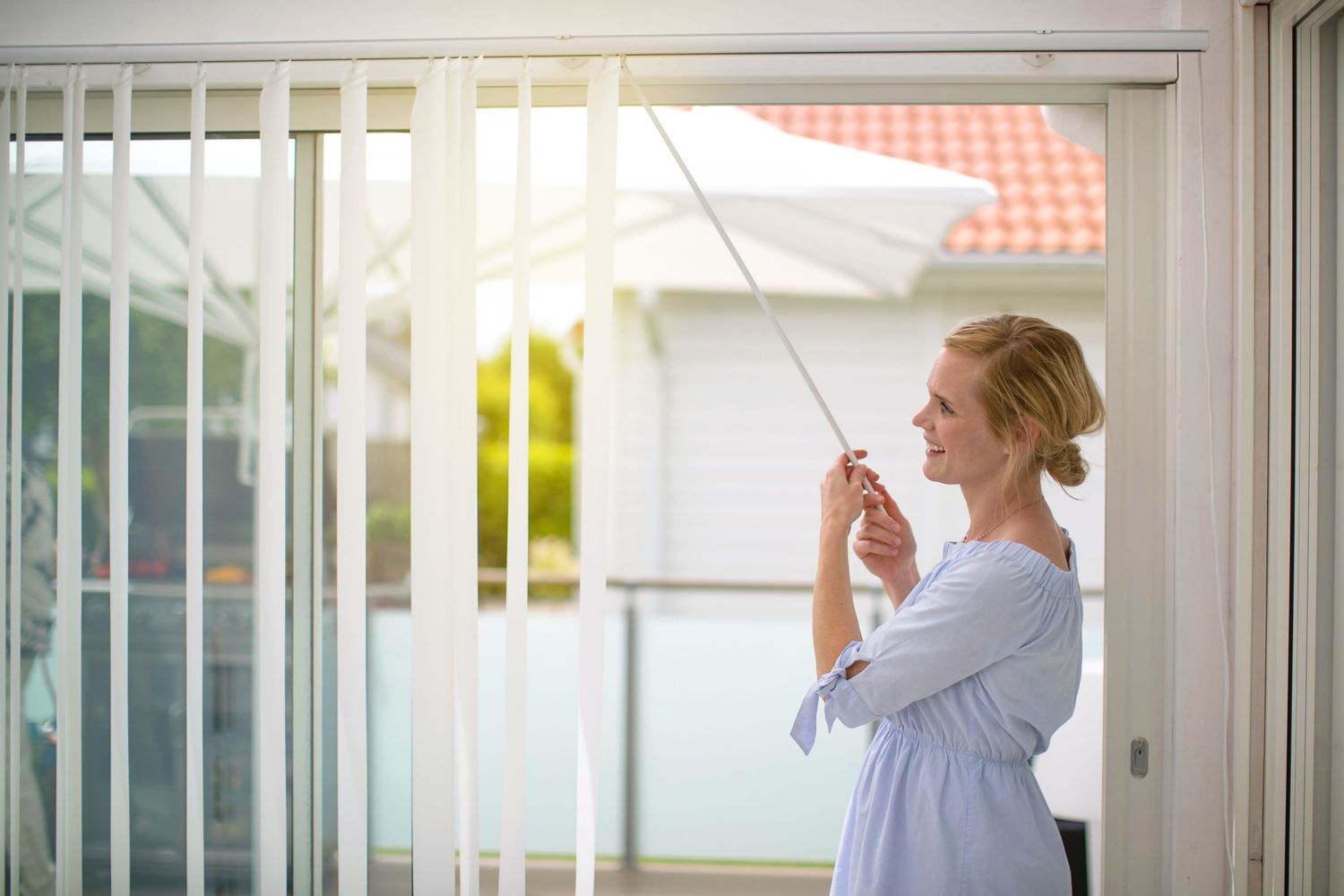 Woman in white dress drying laundry by window with sunlight streaming through sheer curtains.