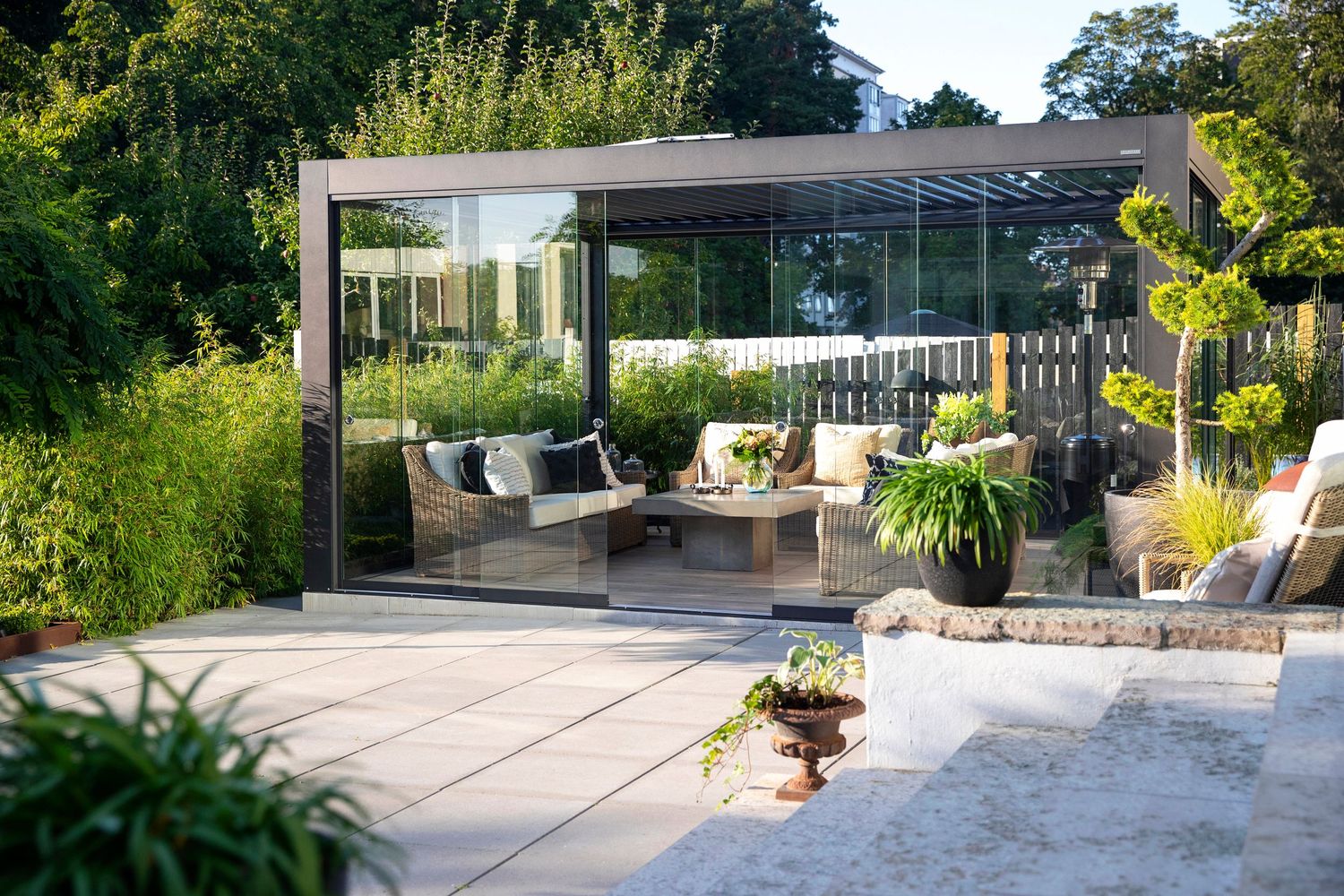 Modern outdoor patio with glass pergola, comfortable seating, and lush garden surroundings.