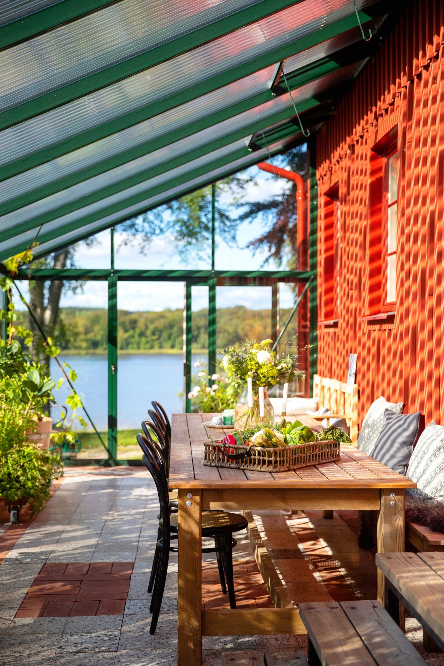 Sunlit lakeside veranda with wooden furniture and green-roofed glass panels.