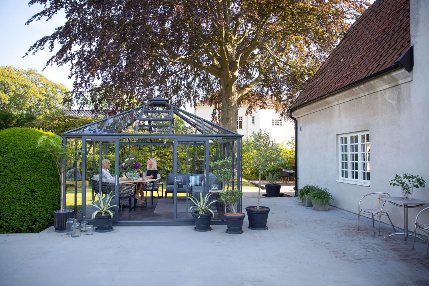 Elegant glass greenhouse with potted plants in courtyard of white building.