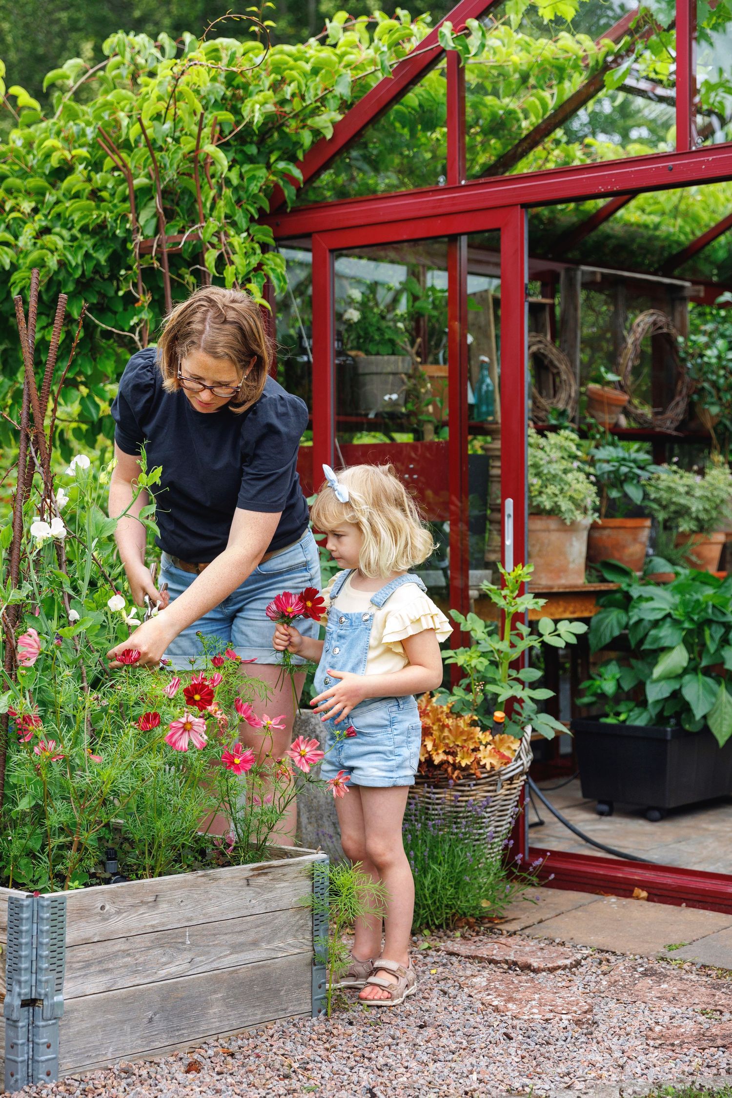 Adult and child tending to flowers in raised garden bed near red greenhouse structure.