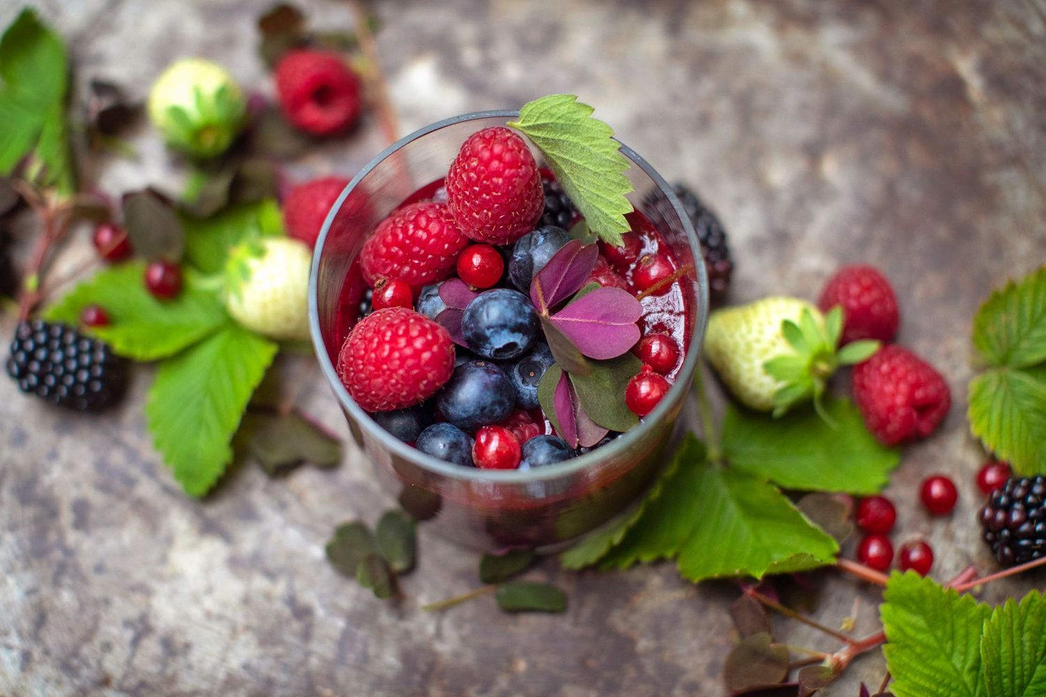 Fresh mixed berries and fruits artfully arranged in a glass with green leaves.