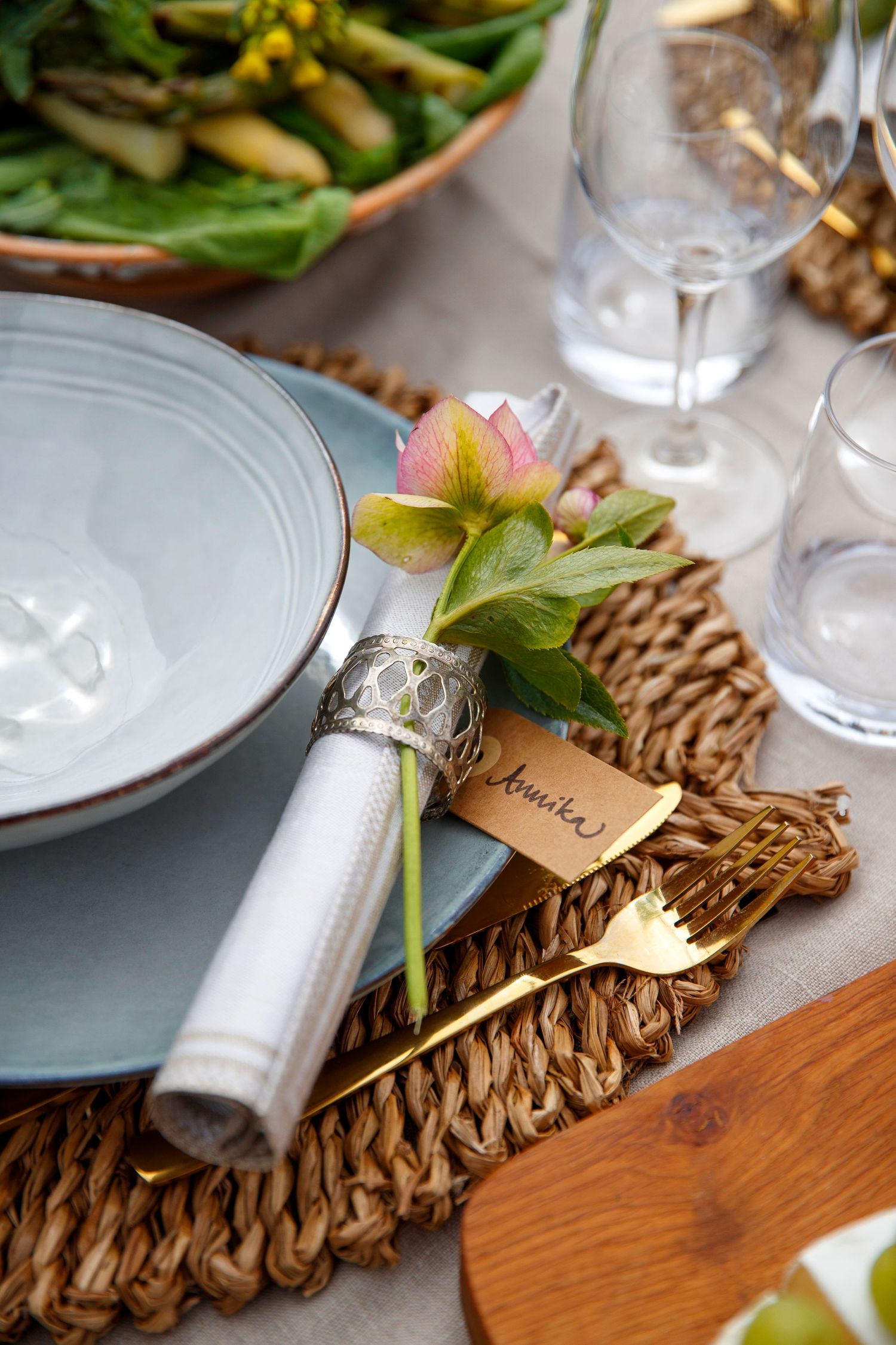 Elegant table setting with woven placemats, white napkins, brass cutlery and delicate place cards on wooden table.