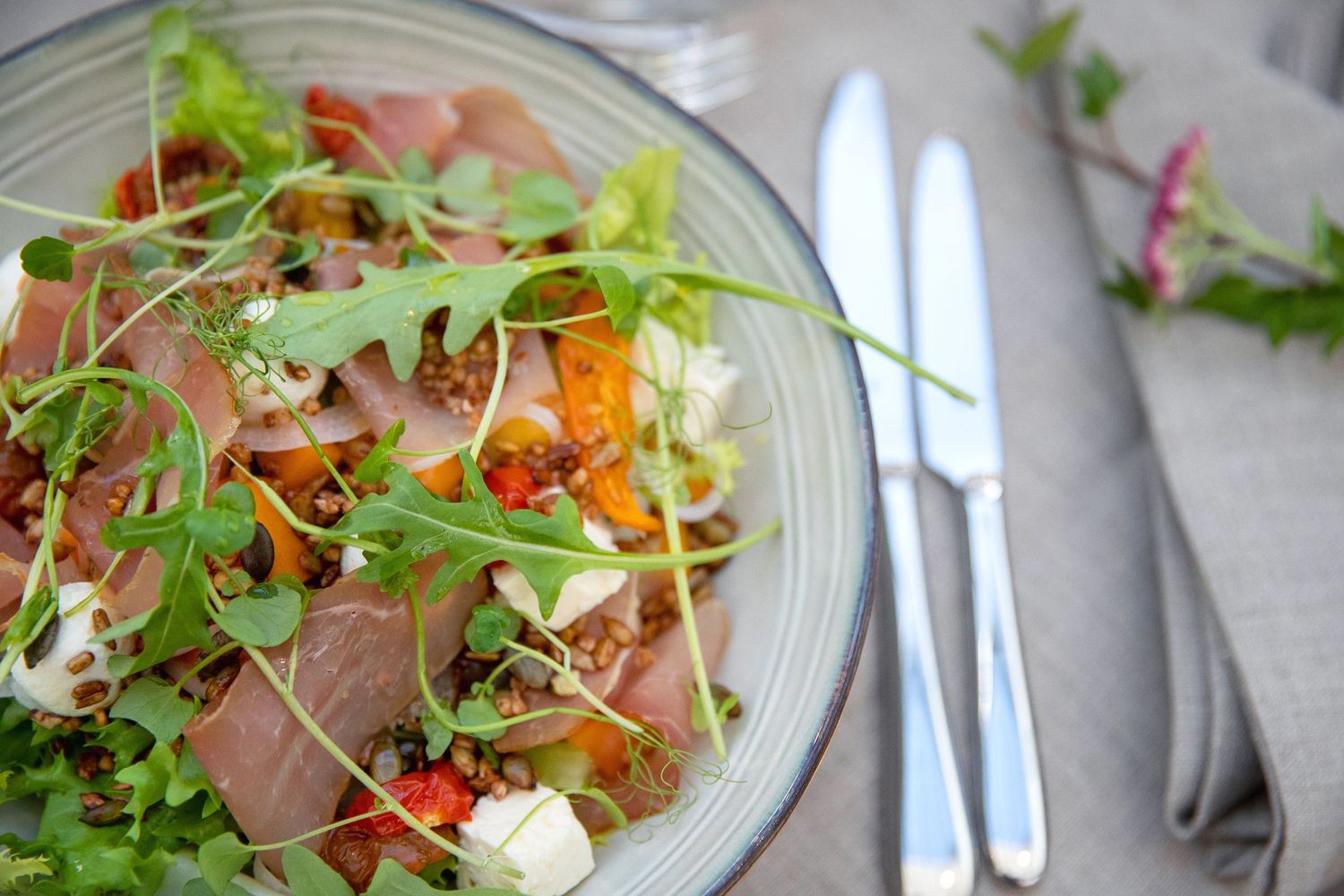 Fresh salad bowl with mixed greens, prosciutto, and colorful vegetables served with cutlery.