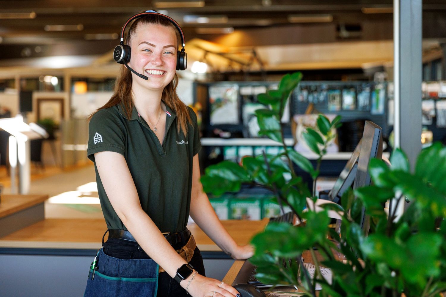 Customer service representative wearing headset smiles at service desk with plants.