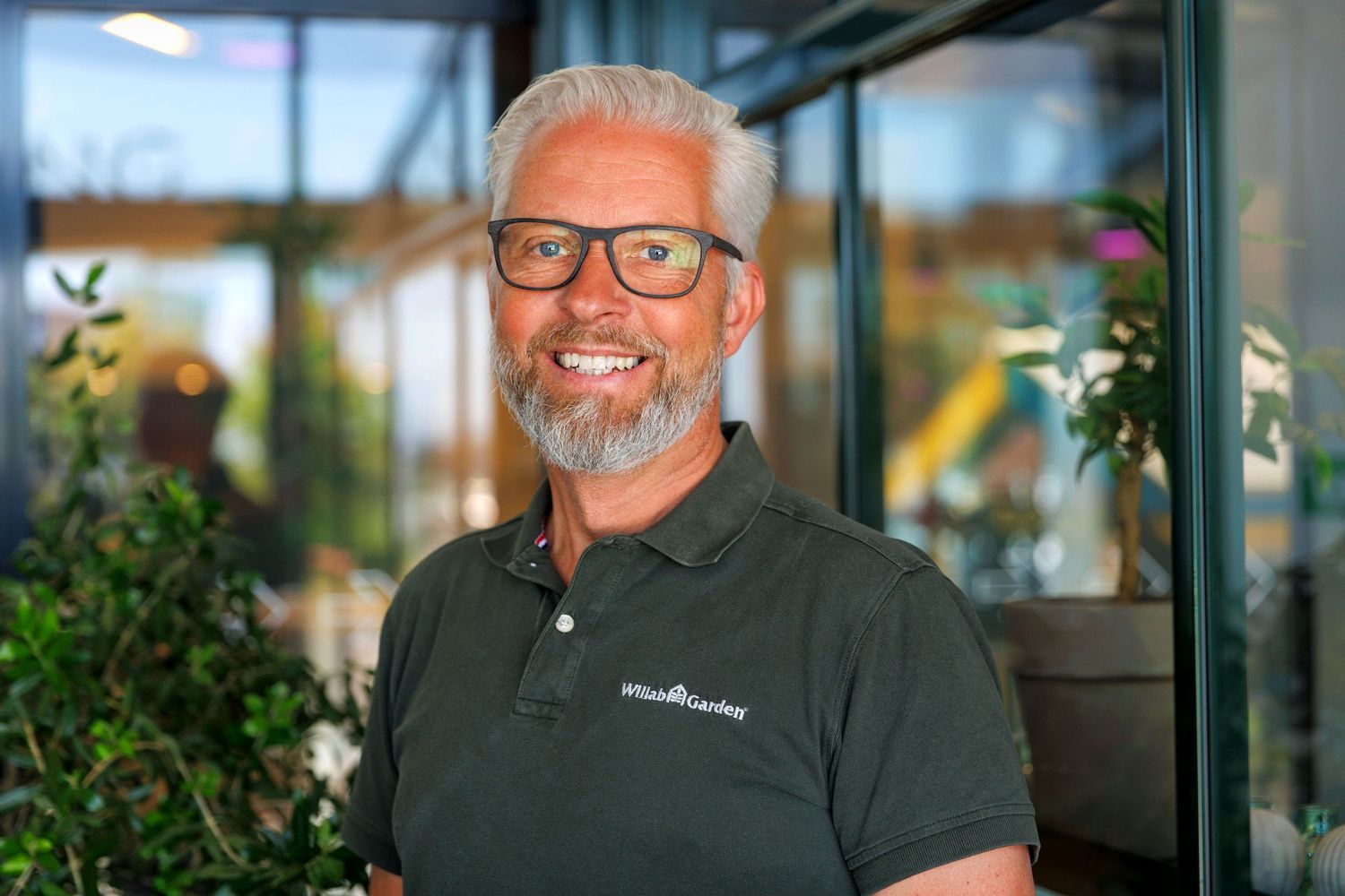 Professional in glasses and dark polo shirt standing in front of greenhouse windows.