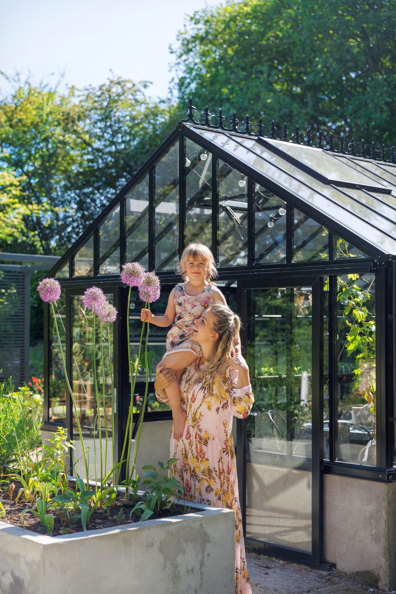 Adult and child in floral dresses in front of a black-framed greenhouse in a sunny garden.