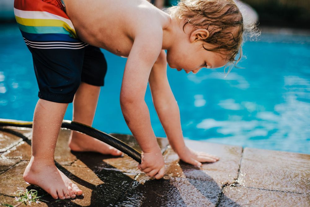 Young child in rainbow swim trunks bends down to touch water at edge of bright blue swimming pool on sunny day.