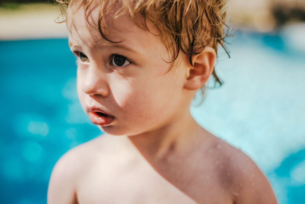 Small child with wet curly hair gazes sideways against bright blue swimming pool background.
