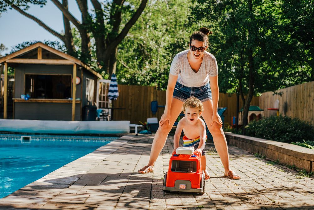 A person in sunglasses and shorts stands on a poolside patio with a bright red toy car on a sunny summer day.