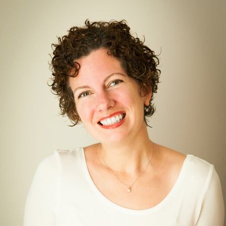 A headshot portrait of someone with curly dark hair wearing a white top against a neutral background.