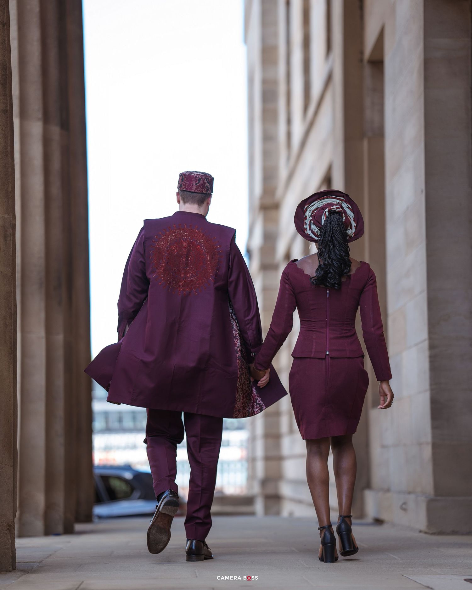 Stylish couple wearing coordinated burgundy outfits walking together between elegant stone columns.