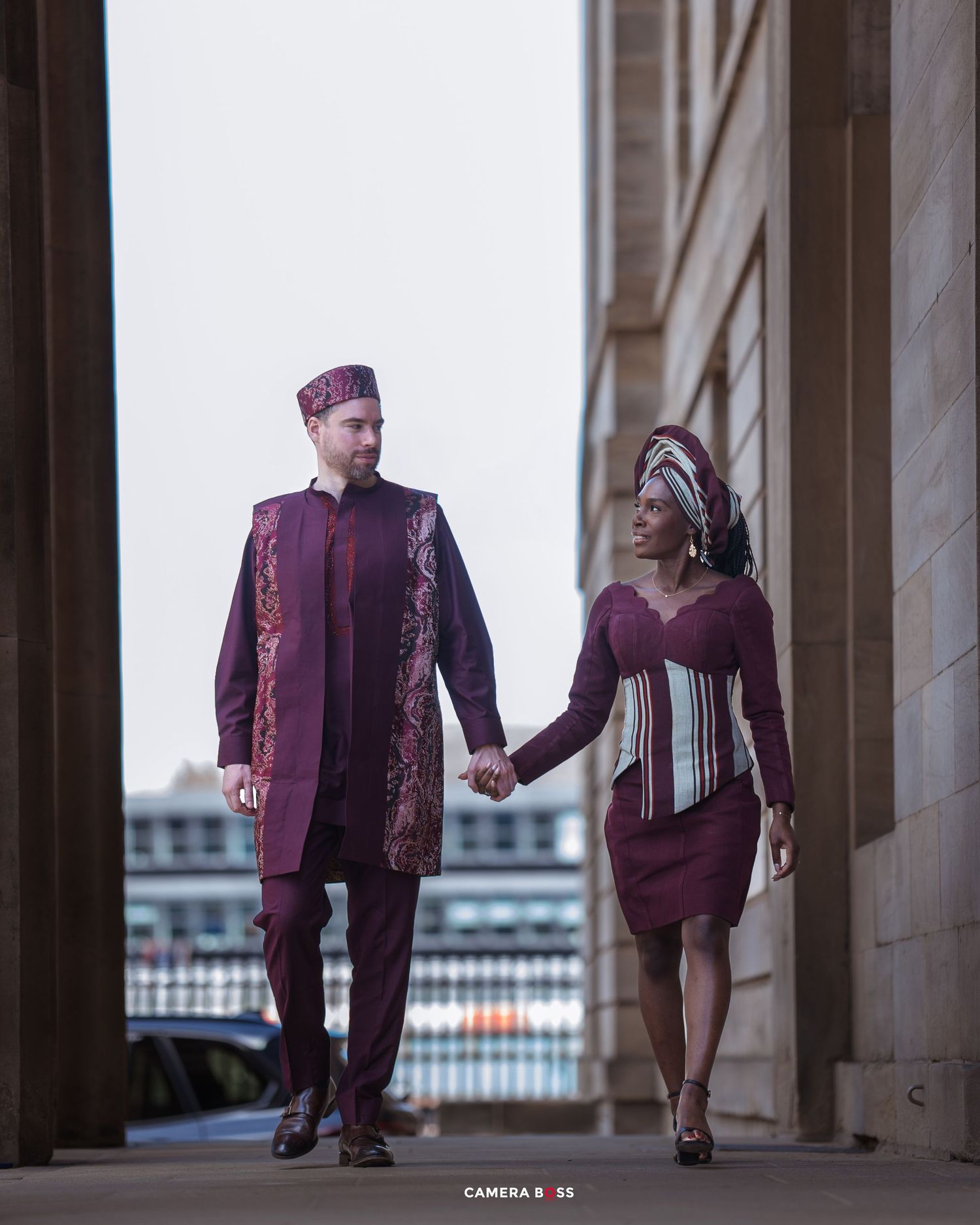 A couple in coordinated burgundy formal attire poses in an elegant covered walkway with stone columns.