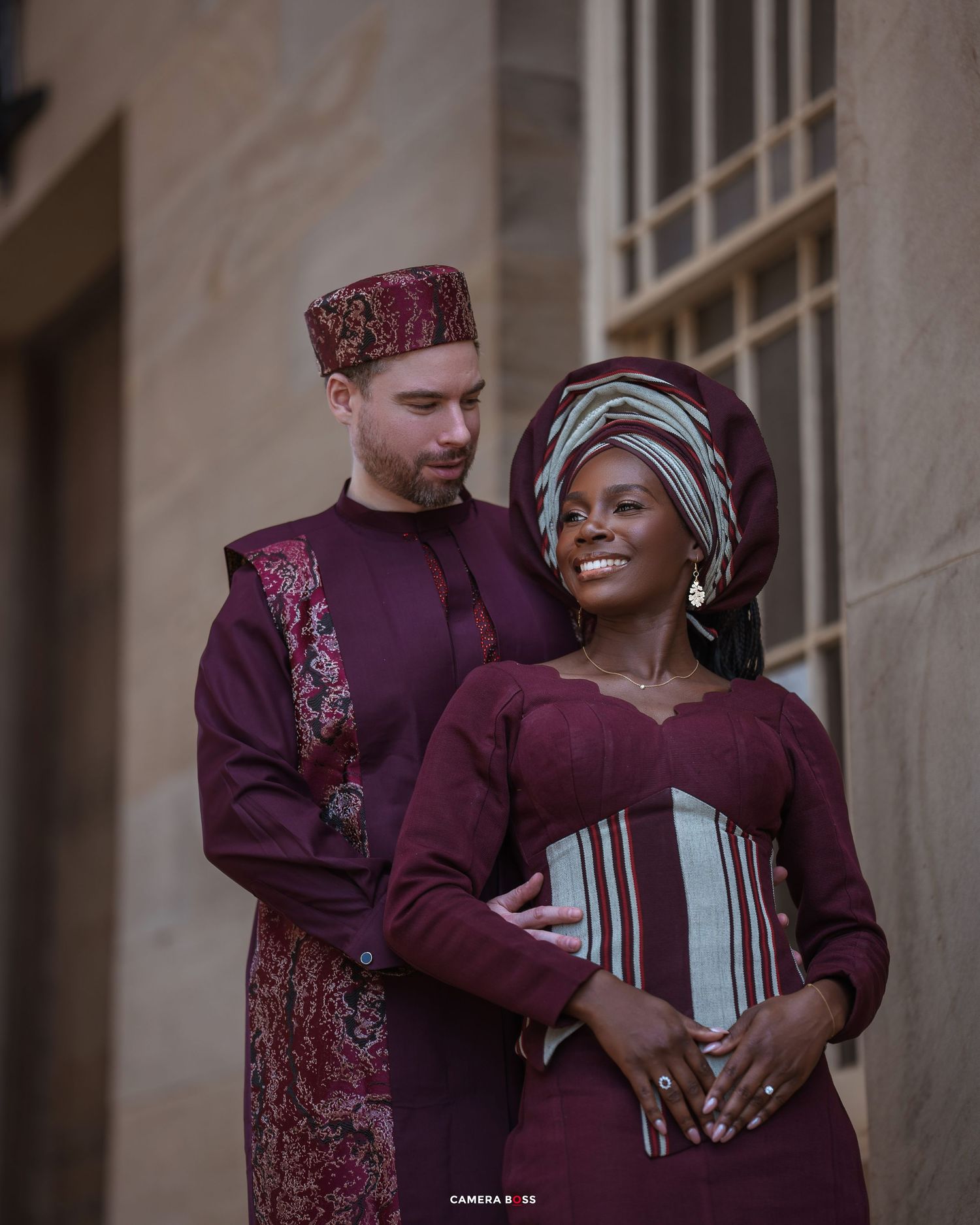 A couple in coordinating burgundy traditional attire pose together, with her wearing a gele headwrap and him in ethnic formal wear.