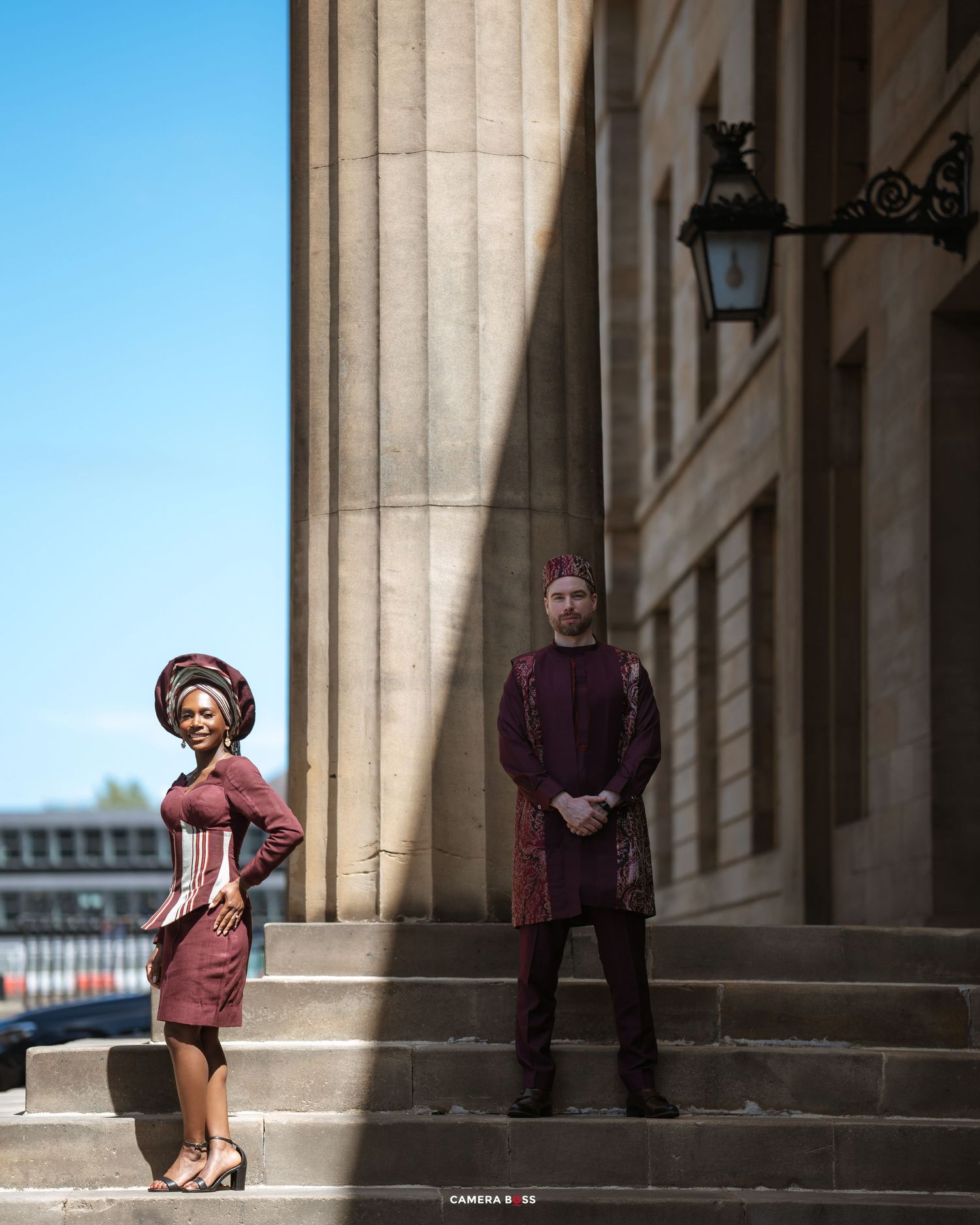 Two people in period costumes pose on stone steps by classical columns, with one in a peach dress and another in purple robes.