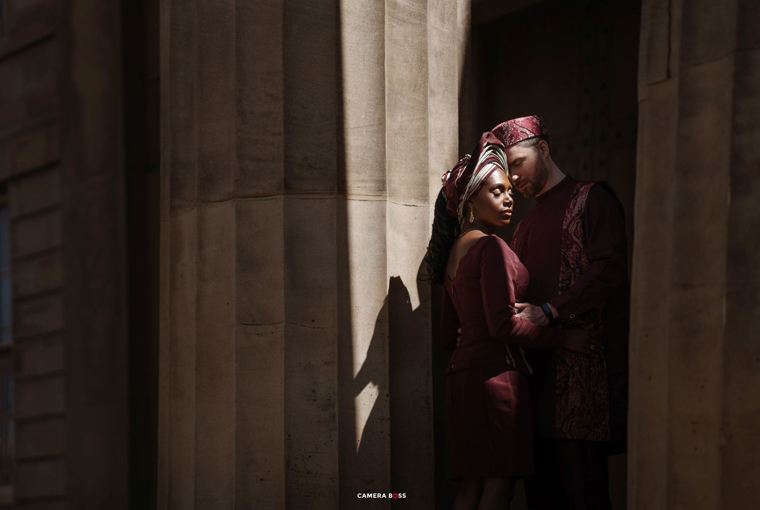A person in maroon clothing stands in shadows between stone columns with dramatic lighting.
