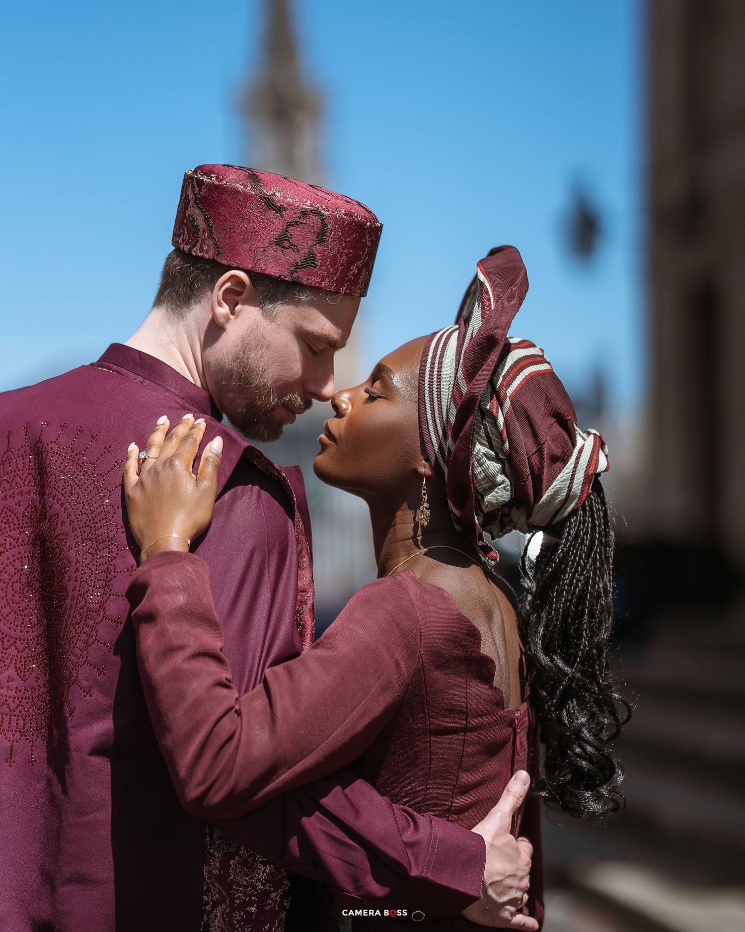 A romantic couple in matching burgundy traditional attire embrace intimately against a bright blue sky.