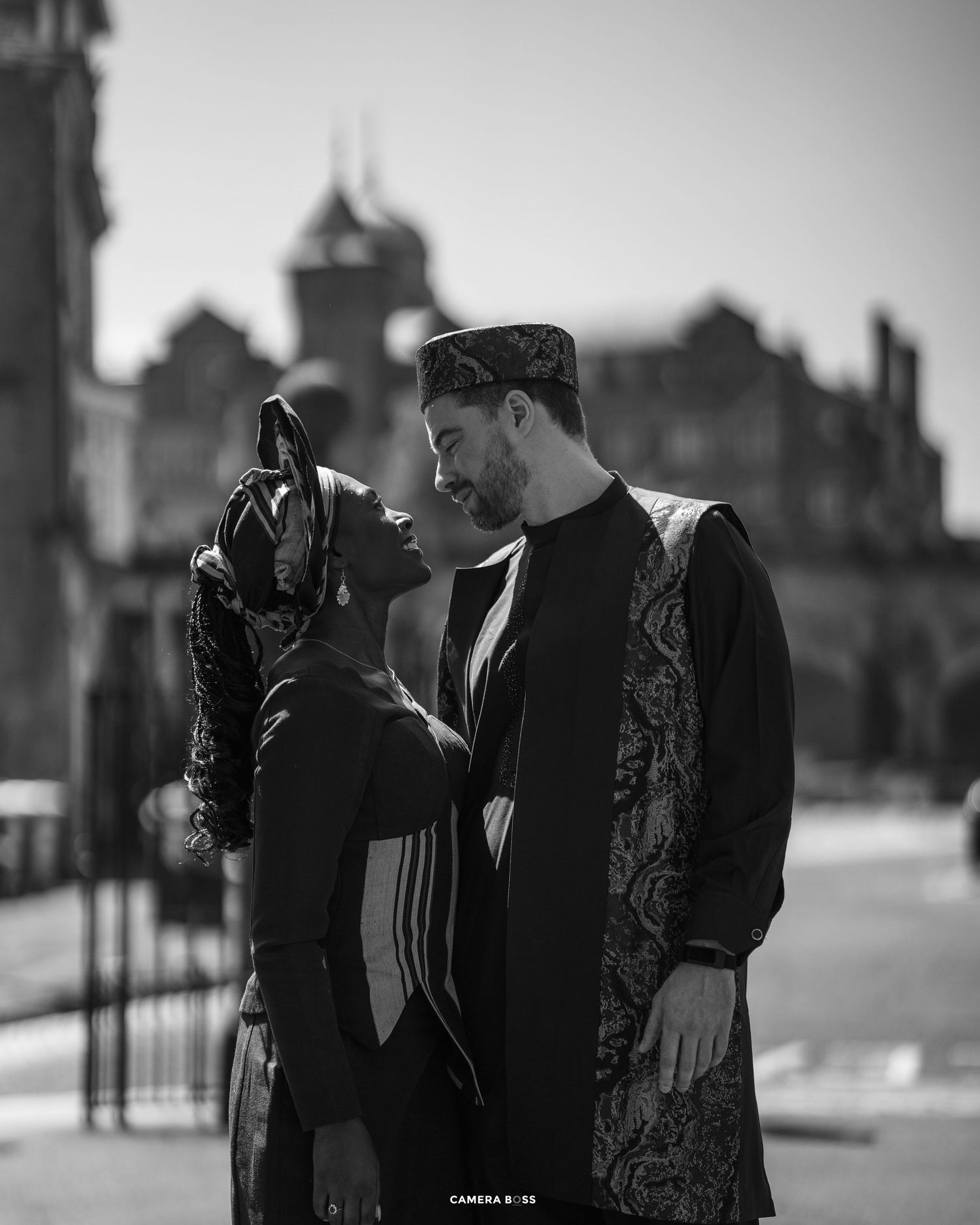 A romantic black and white portrait of a fashionable couple in traditional outfits standing before historic architecture.