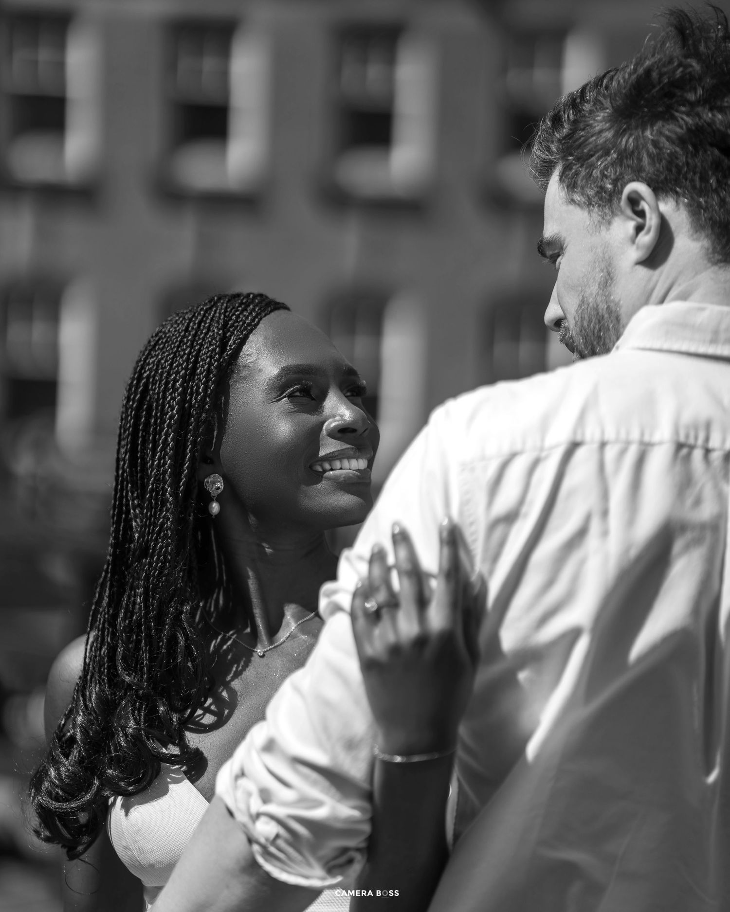 A romantic black and white photo of a couple gazing at each other with gentle smiles against a blurred urban backdrop.