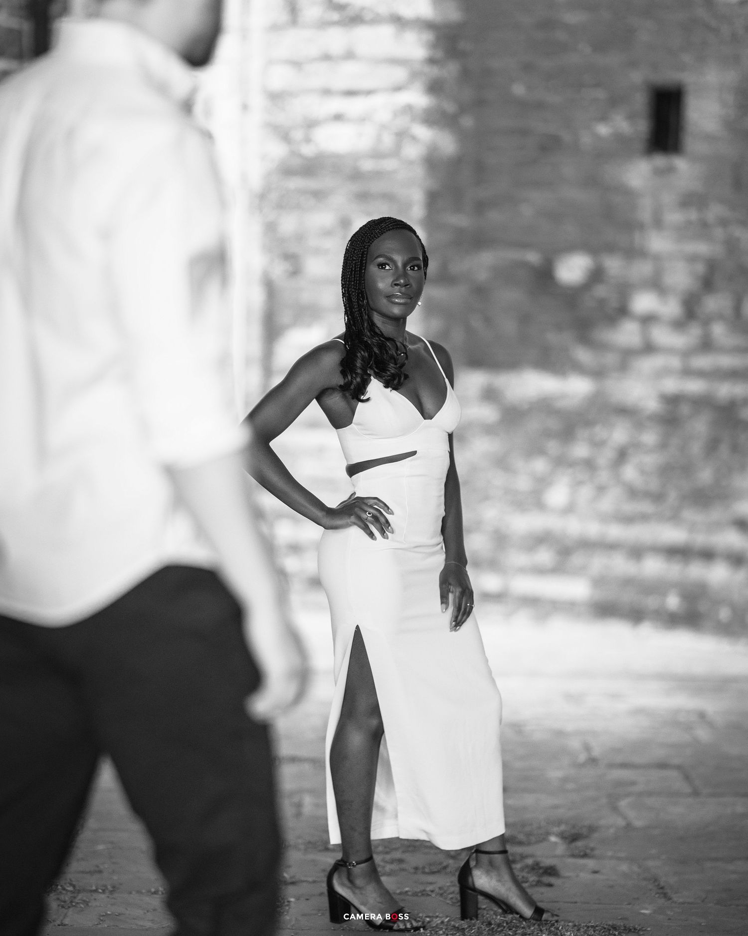 Black and white portrait showing a person in an elegant white dress striking a pose against a textured brick wall background.
