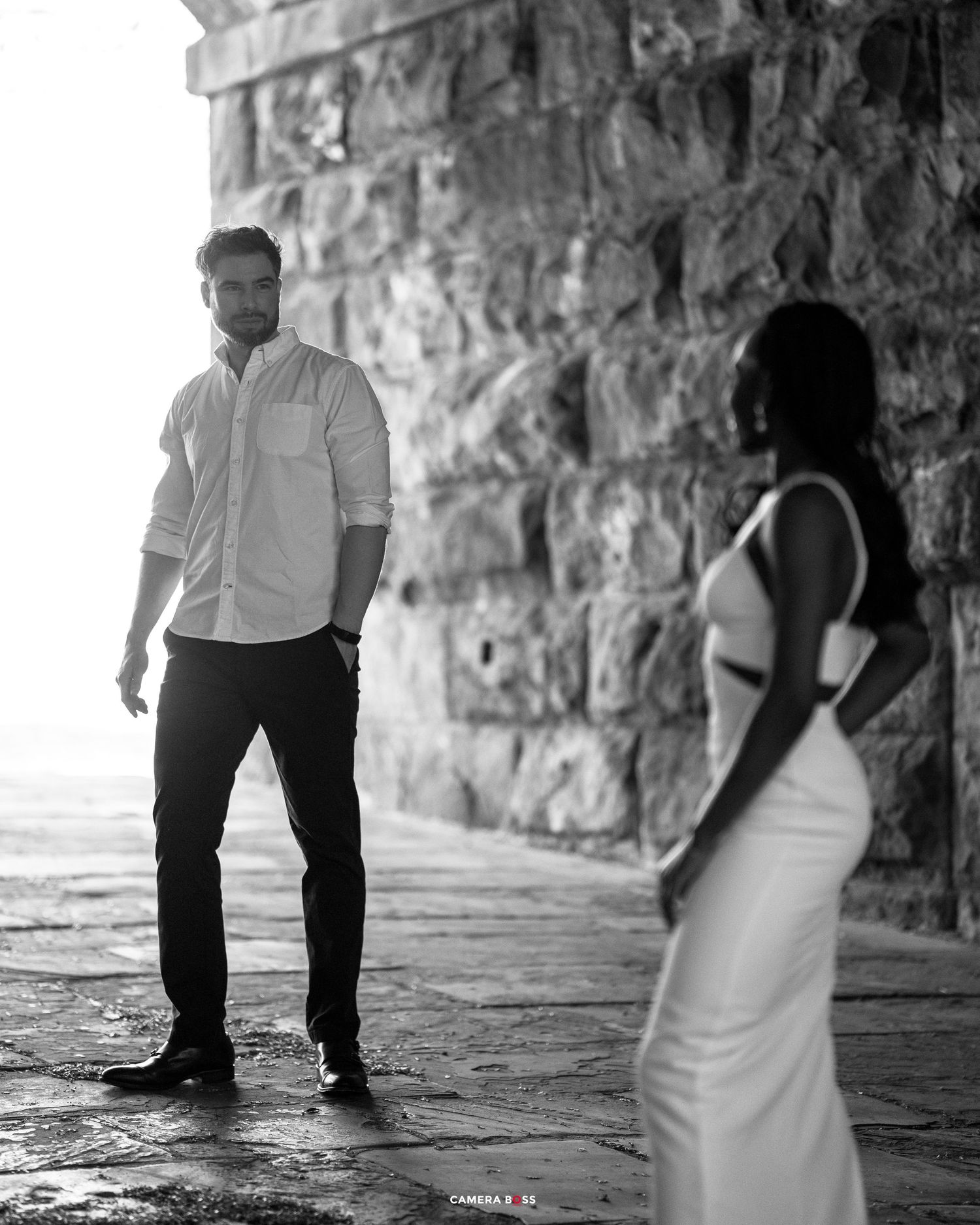 A black and white photo of a couple posing against a stone wall in an artistic engagement photography style.