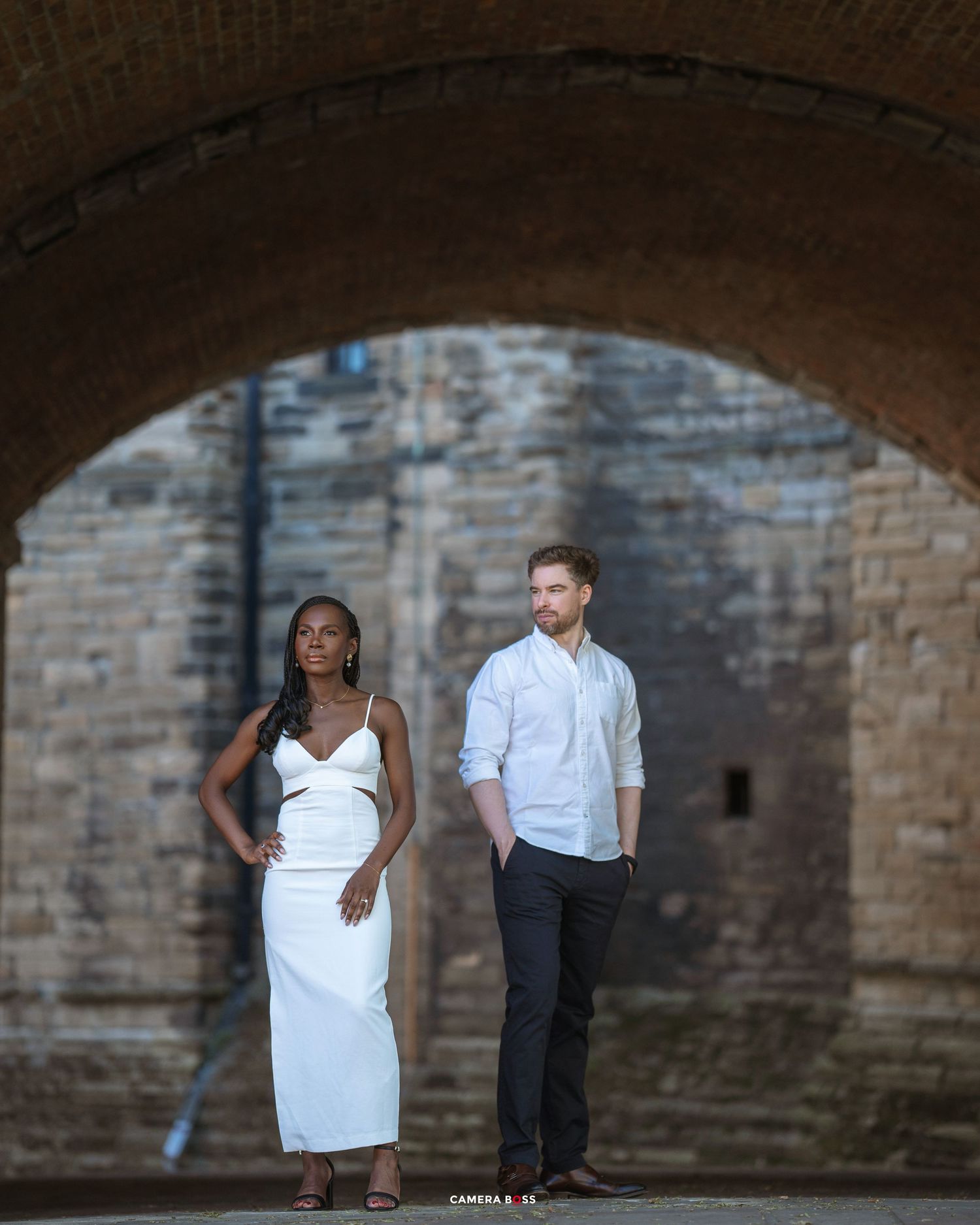 A romantic couple in formal attire poses together under a stone archway on a sunny day.