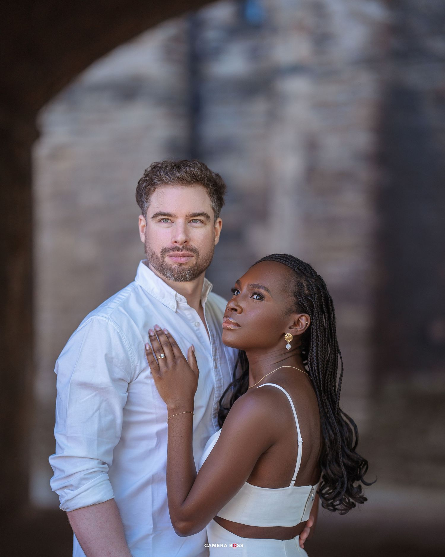 A romantic couple in elegant attire poses intimately against a stone wall backdrop in soft lighting.