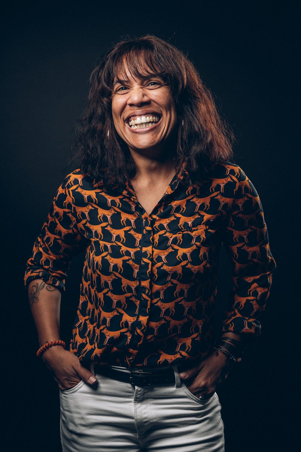 Portrait of someone wearing a patterned orange shirt smiling warmly against a dark background.