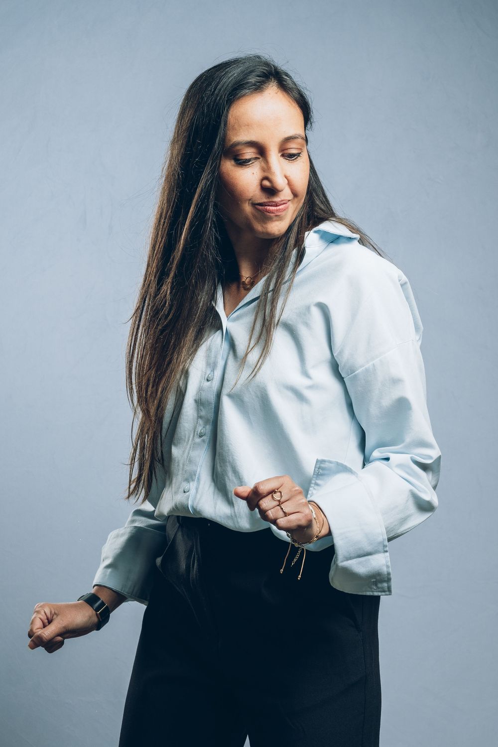 Person wearing a light blue button-down shirt and black pants stands casually against a gray background.