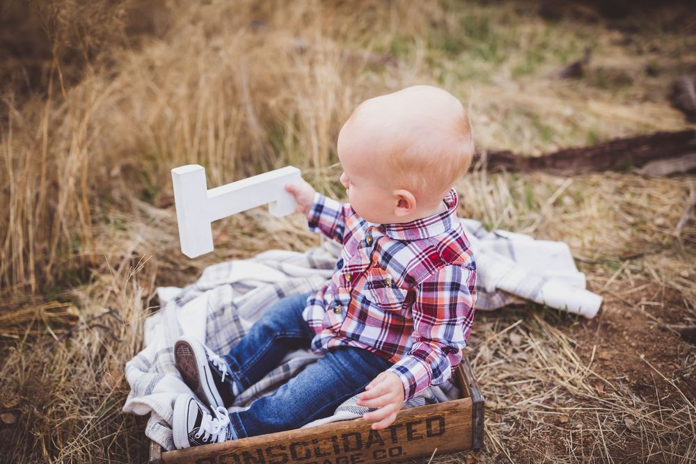 A small child in plaid clothing and blue jeans lies on dried grass during an outdoor photo shoot.