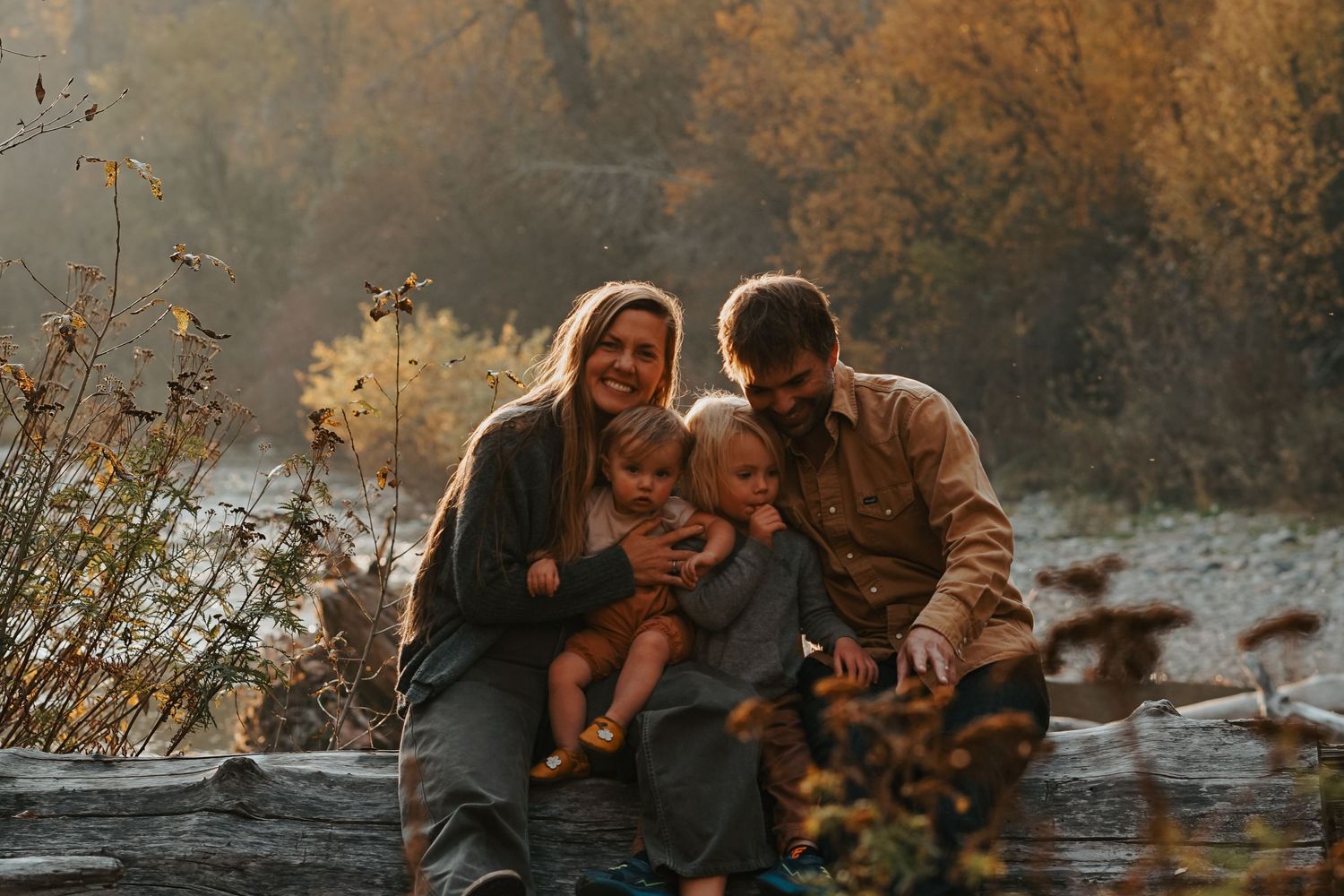 A family sits together on a fallen log by a stream at sunset in an autumn woodland scene.