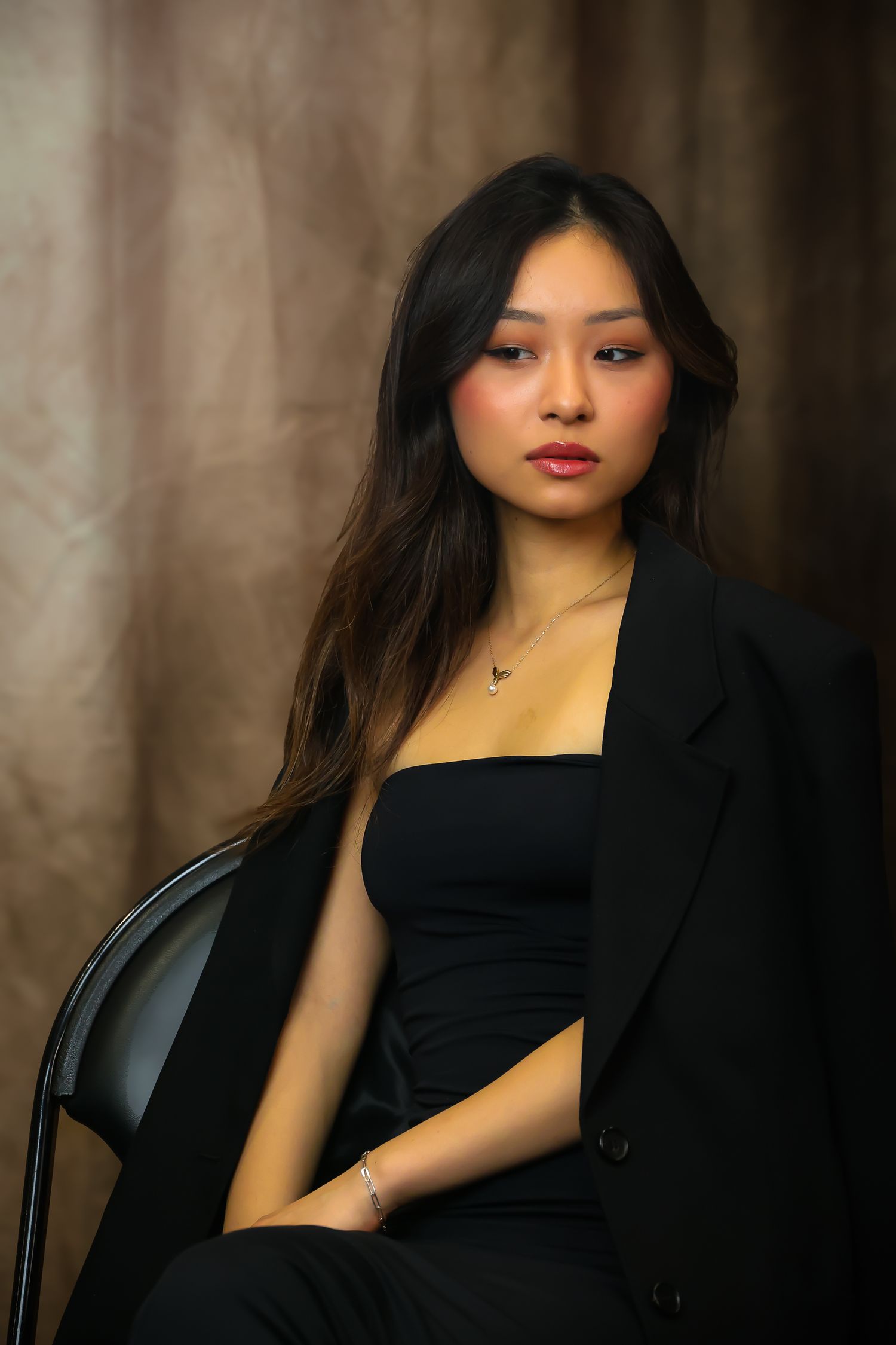 Elegant portrait of someone in a black formal outfit seated against a moody brown background in dramatic lighting.