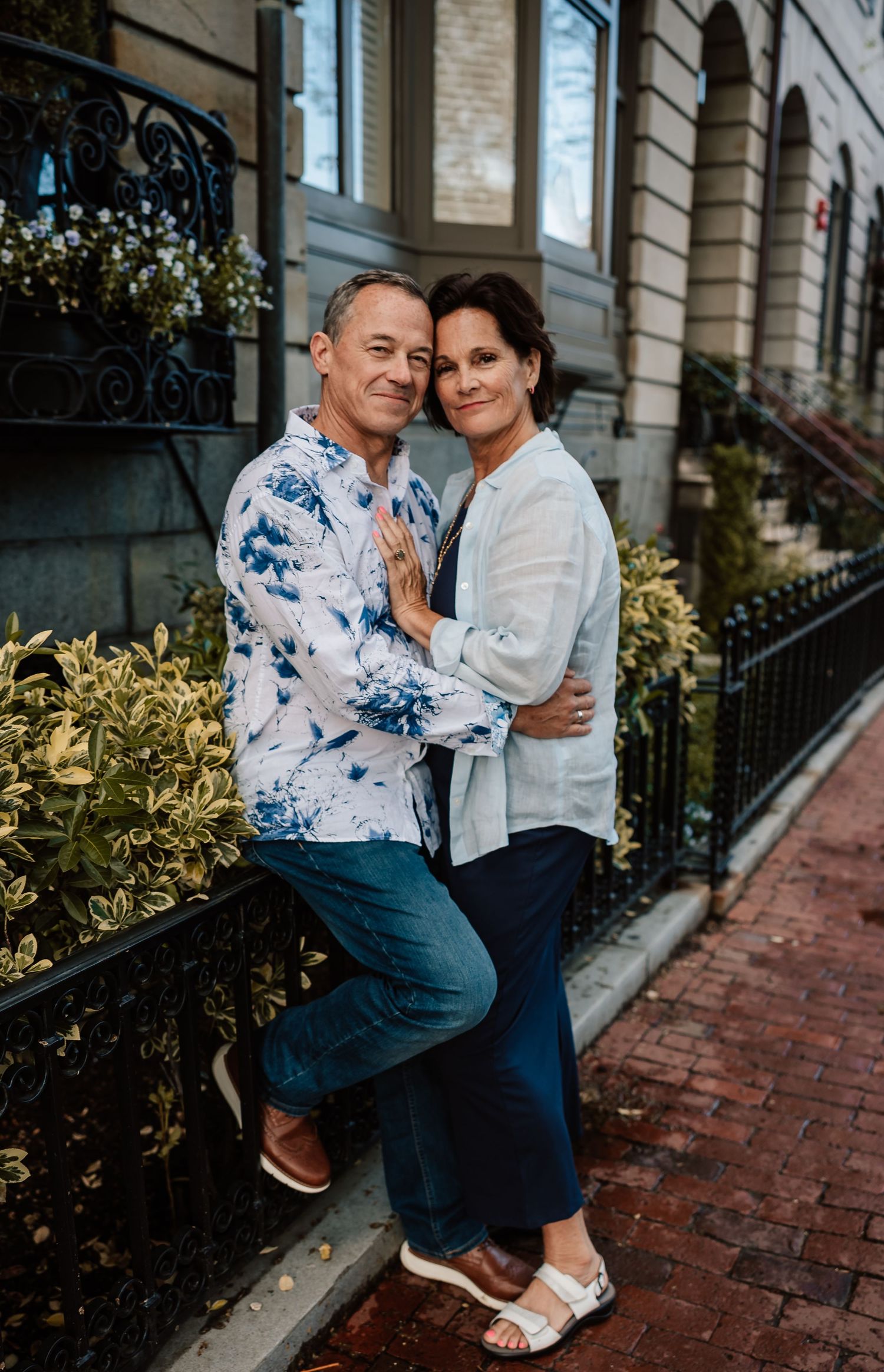 Romantic couple embraces on brick sidewalk beside ornate brownstone buildings in an urban setting.