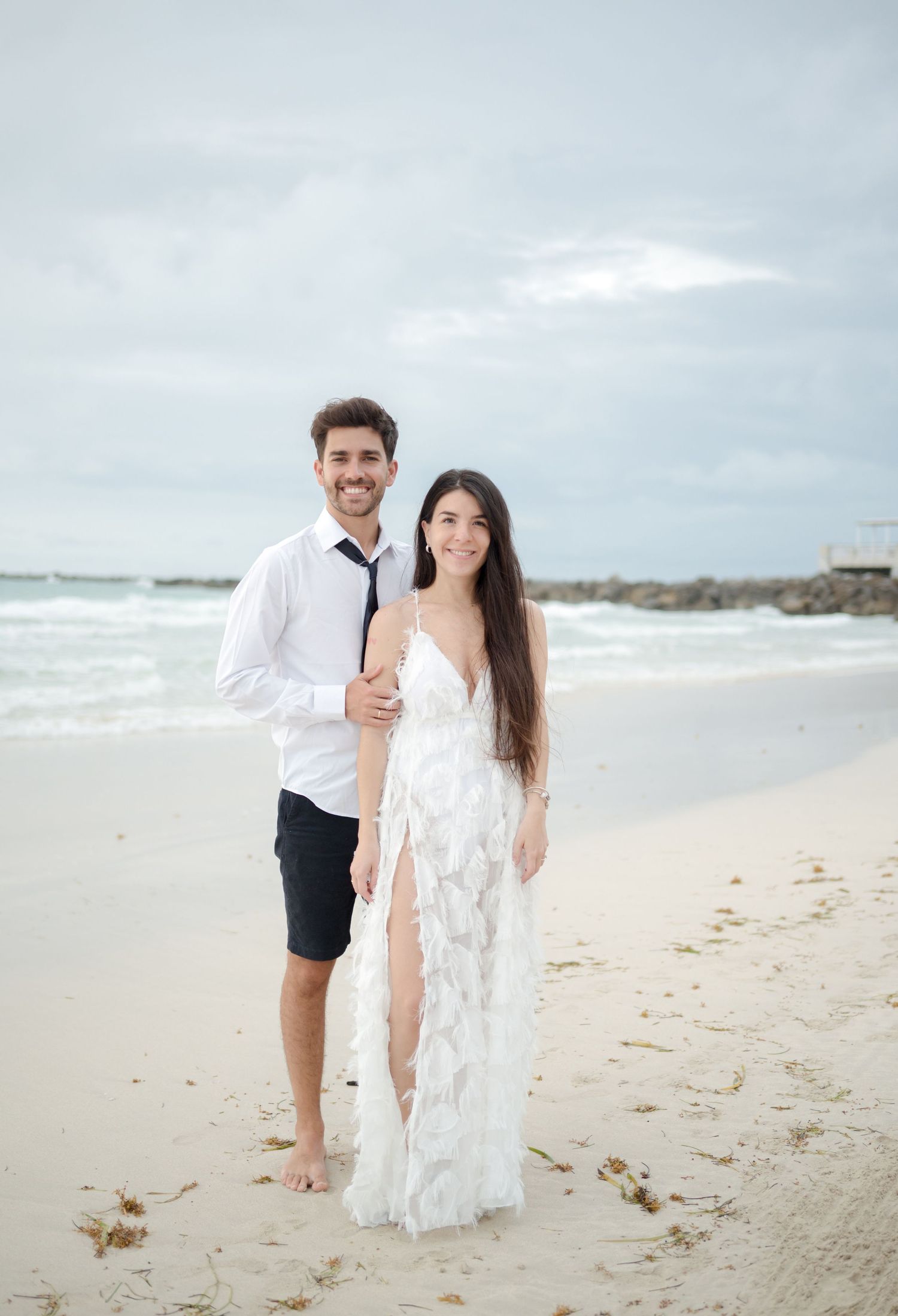 A romantic beach photo at the seashore with a rocky pier in the background on a cloudy day.