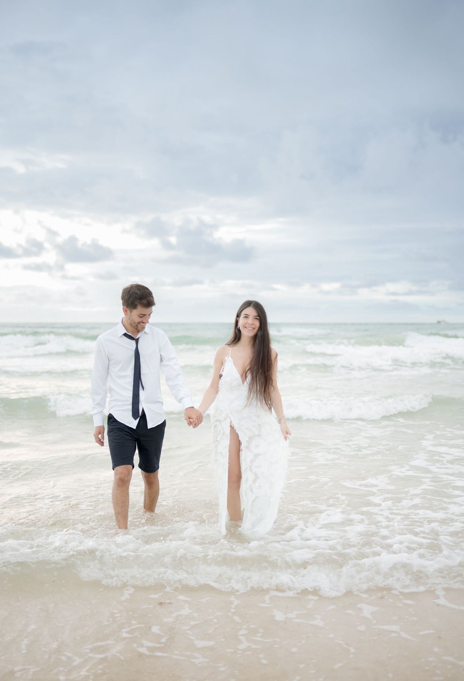 A couple in wedding attire playfully walks hand in hand along the ocean shore during a cloudy beach photo session.
