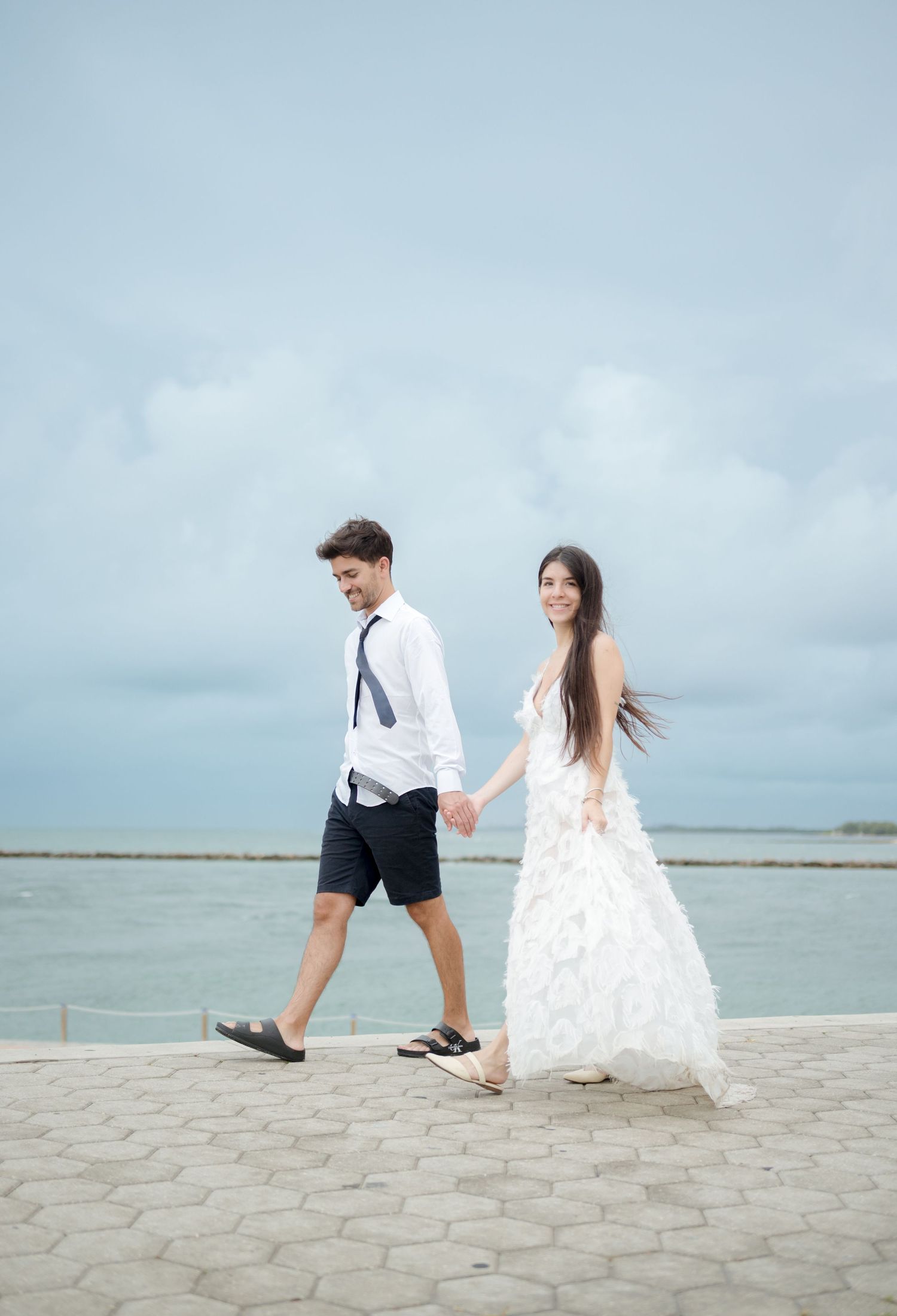 A couple runs playfully together along a coastal walkway with overcast skies in the background.