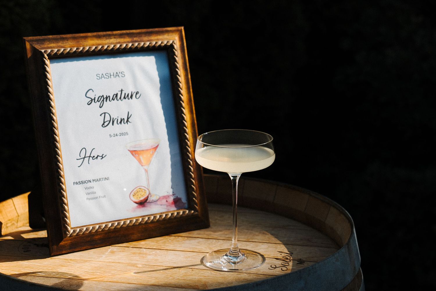 Cocktail glass with creamy liquid next to a framed menu sign on wooden surface in warm lighting.