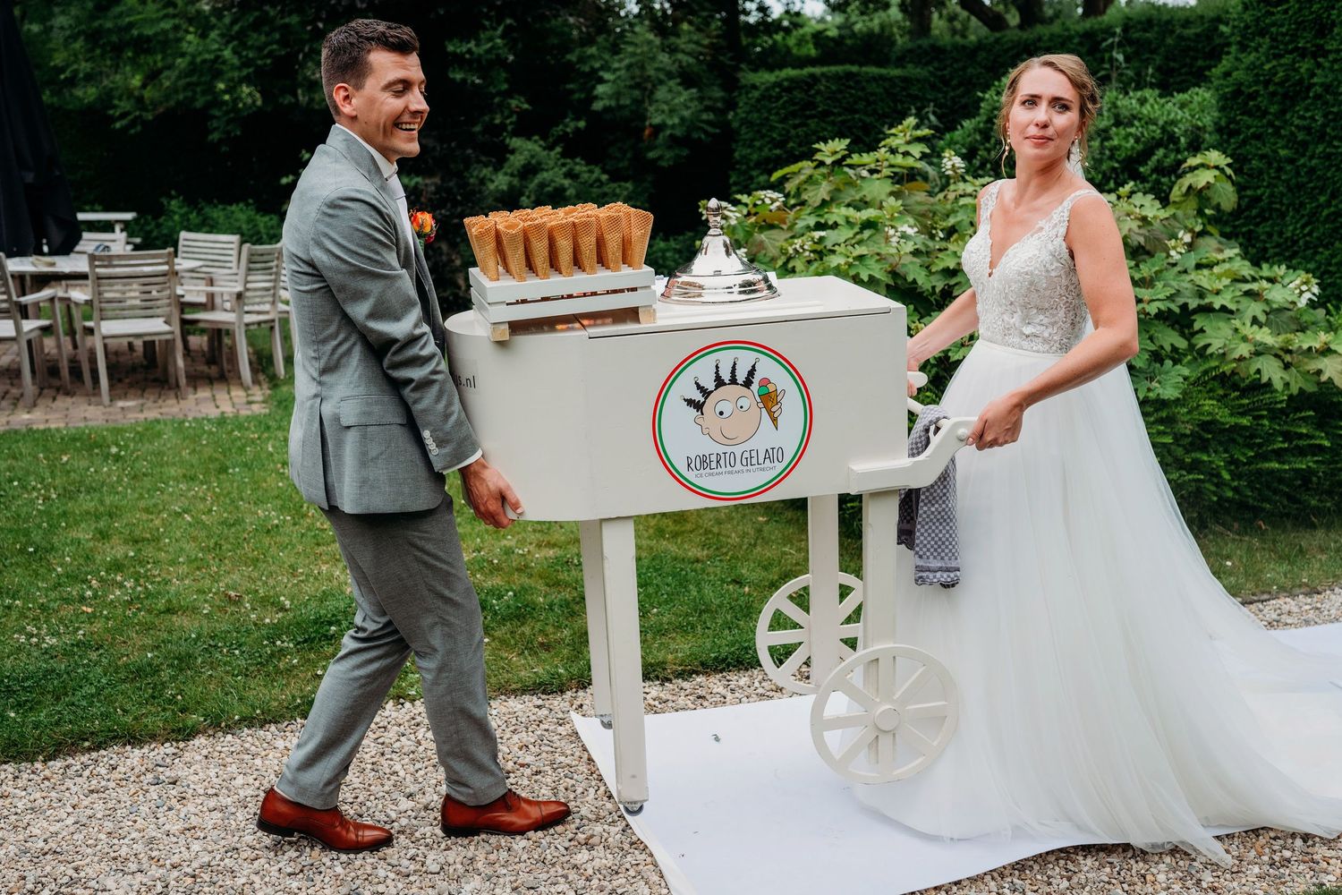A couple poses at a rustic outdoor wedding venue with a decorative white drink cart in a garden setting.