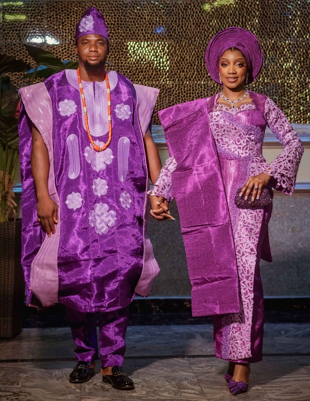 Two people wearing elegant purple traditional Nigerian attire with matching headpieces stand together at formal event.