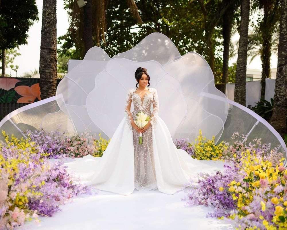 A golden-hour wedding photo with figures standing among vibrant purple and yellow flowers in a garden setting.