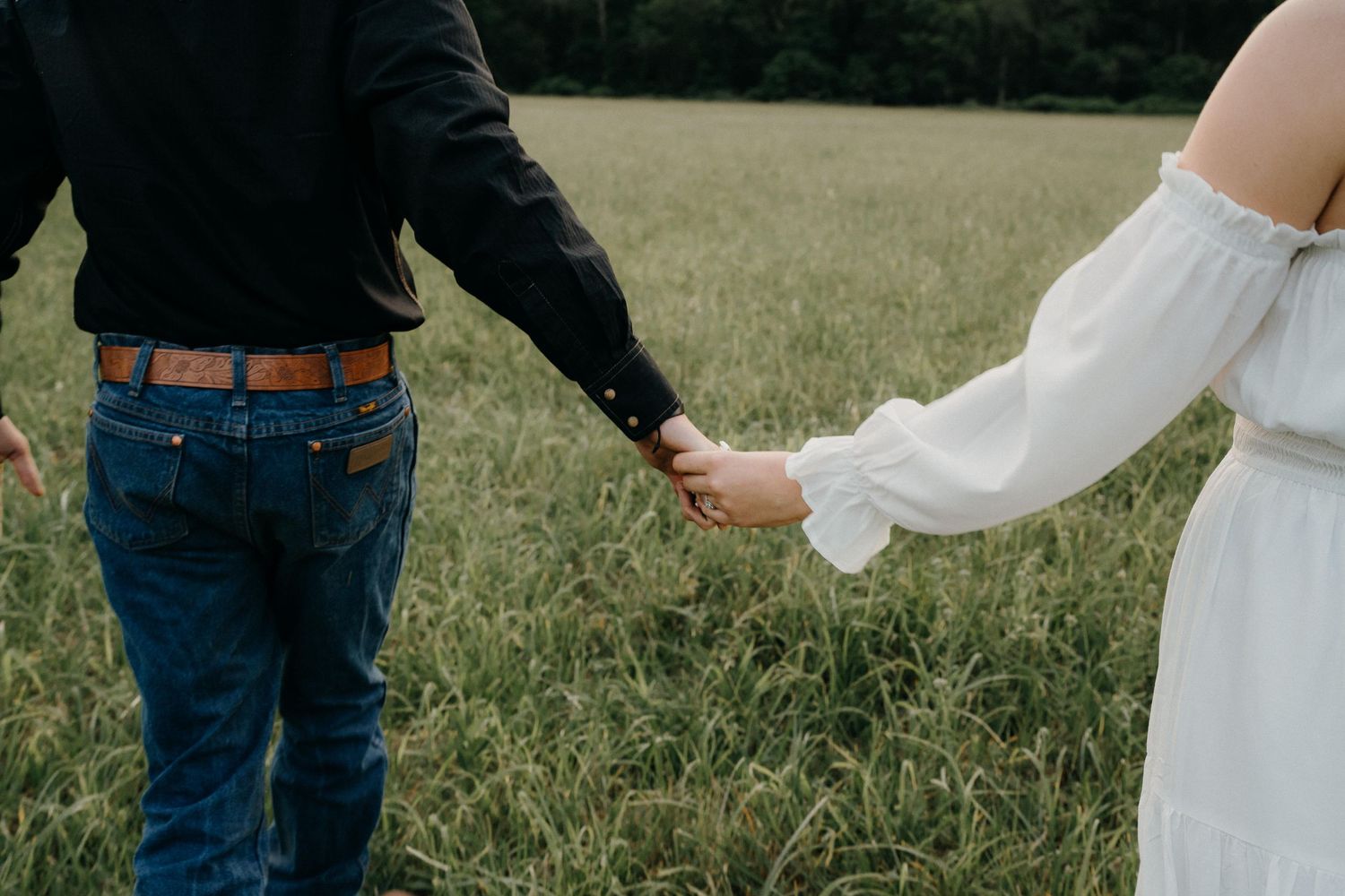 A couple holding hands in a meadow, showing western attire and wedding dress details.